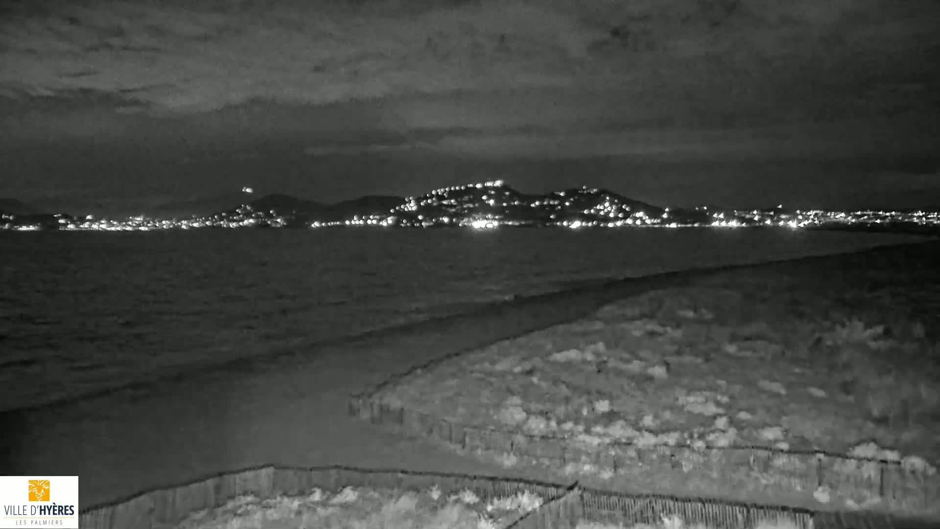 A nighttime view shows a dark beach and calm ocean with a distant, light-studded town nestled against hills under a cloudy sky.
