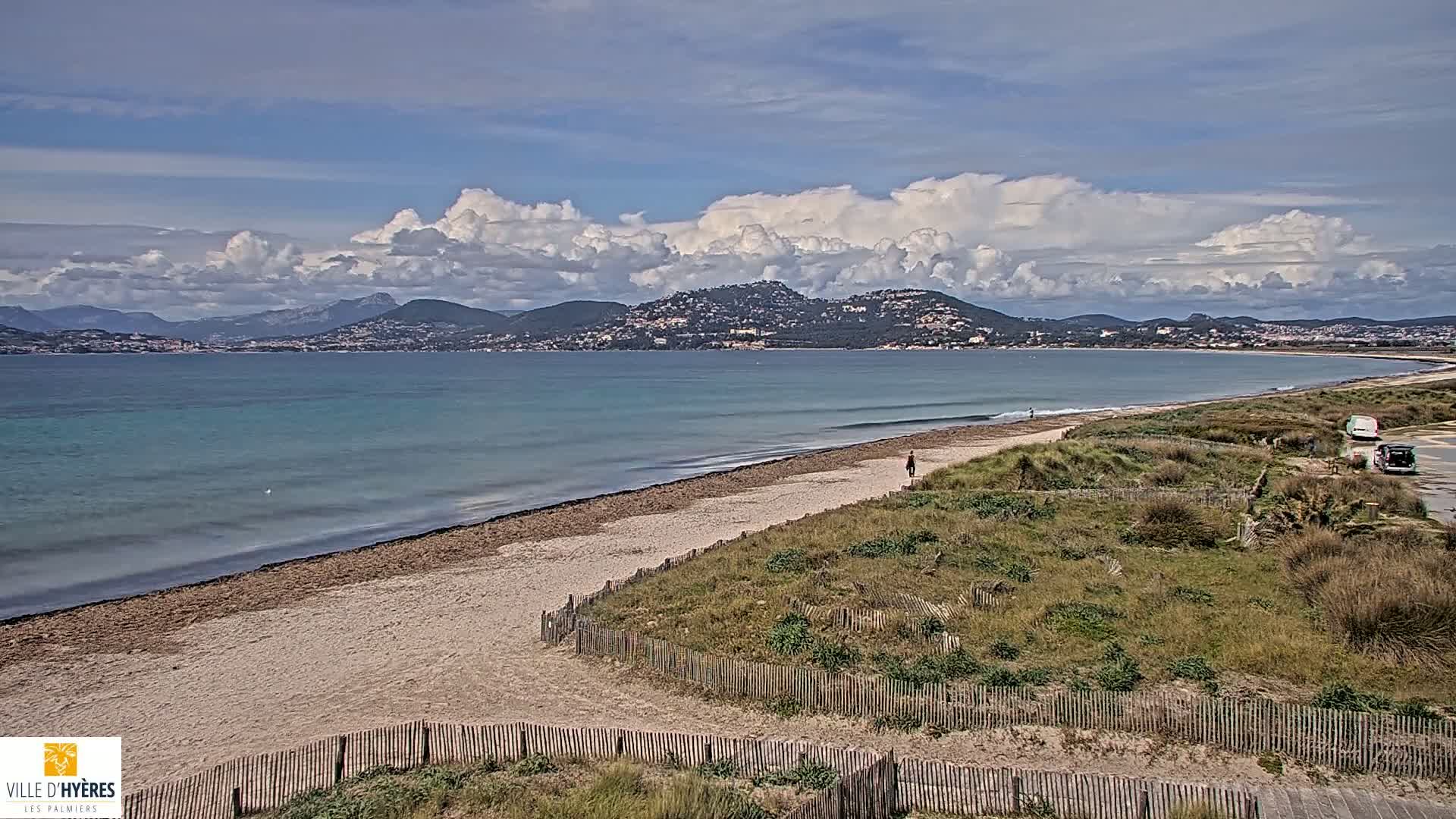 A sandy beach stretches along a calm, teal ocean under a partly cloudy sky, with distant mountains visible in the background.