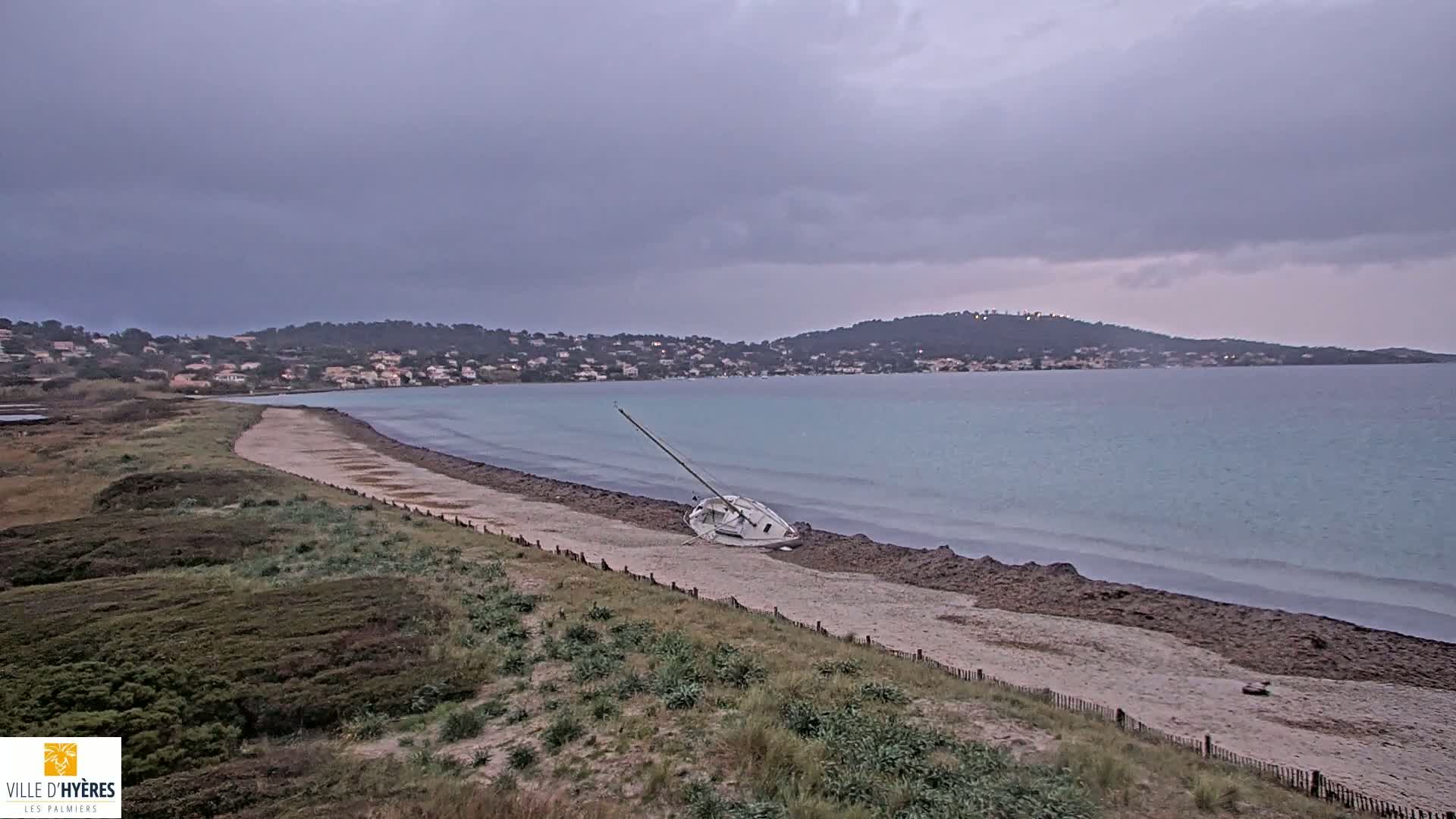 A capsized sailboat lies on a sandy beach under a cloudy, overcast sky, with calm water and a distant town visible across the bay.