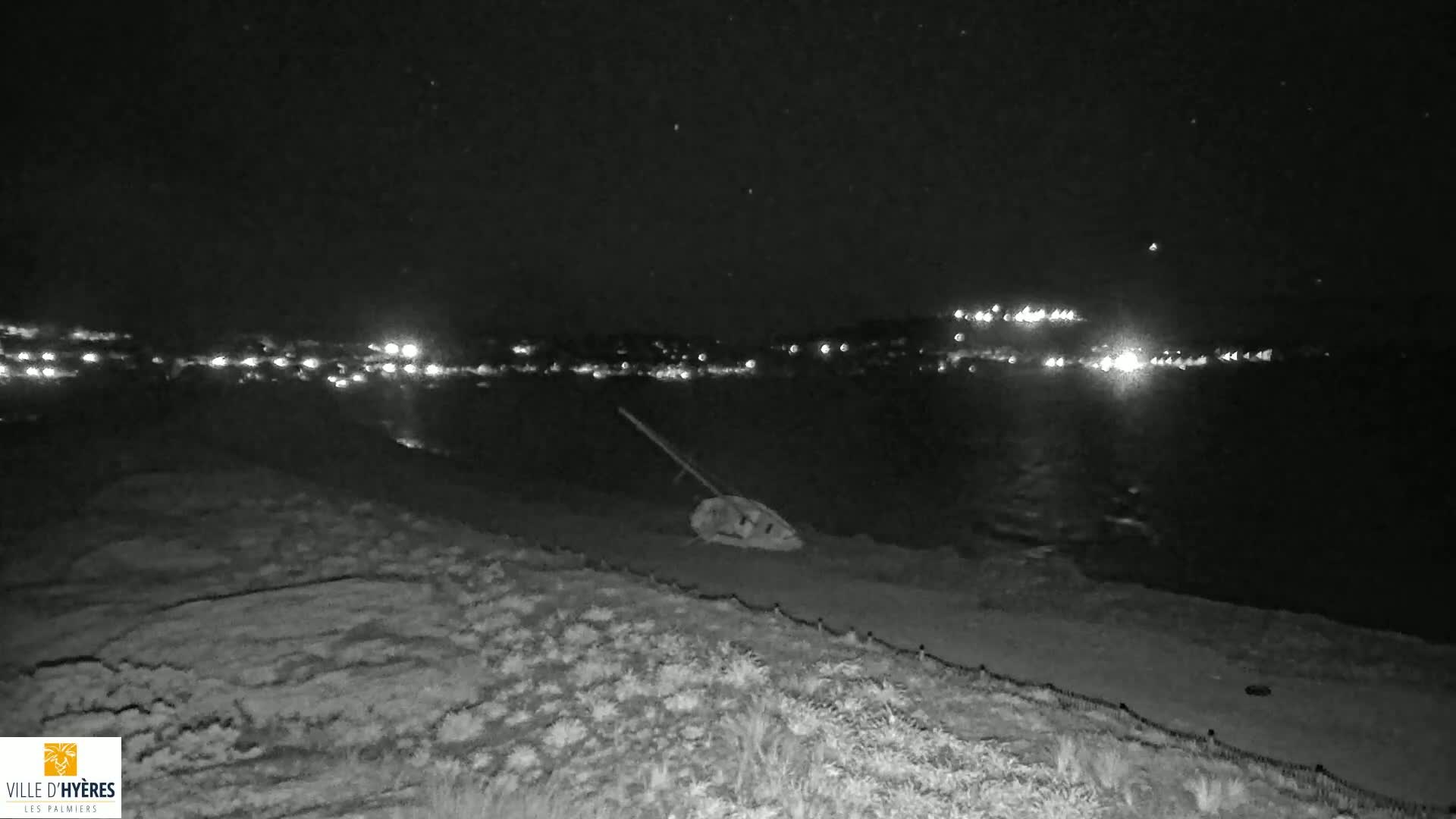 A small boat lies on a dark, sandy shore at night, with a distant town faintly illuminated across the water under a starry sky.