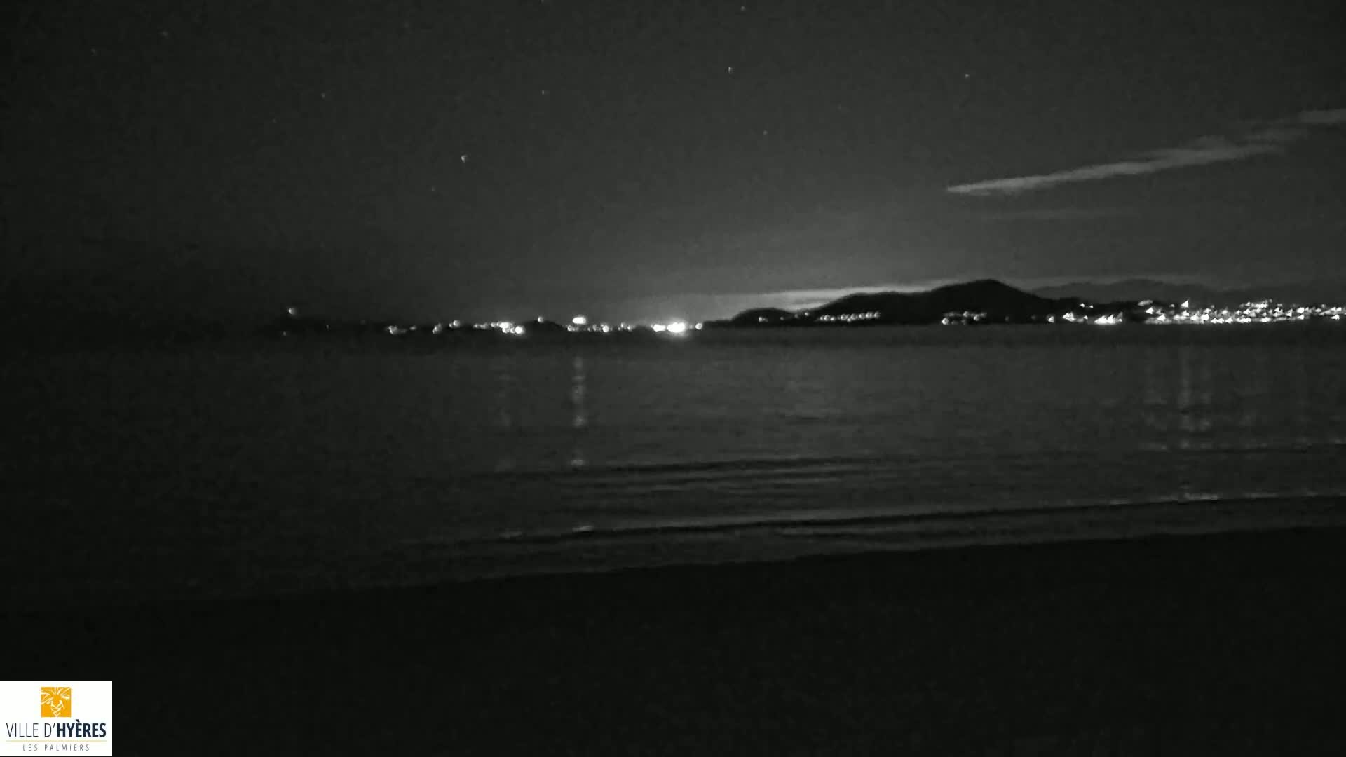 A dark, nighttime beach scene shows a calm sea with a distant city's lights illuminating a dark mountain range under a mostly clear, starry sky.