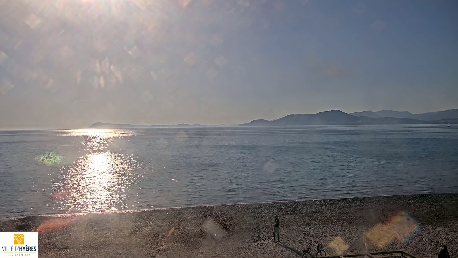 A sunny day reveals a calm sea reflecting the bright sun, with a pebbled beach in the foreground and distant mountains on the horizon.