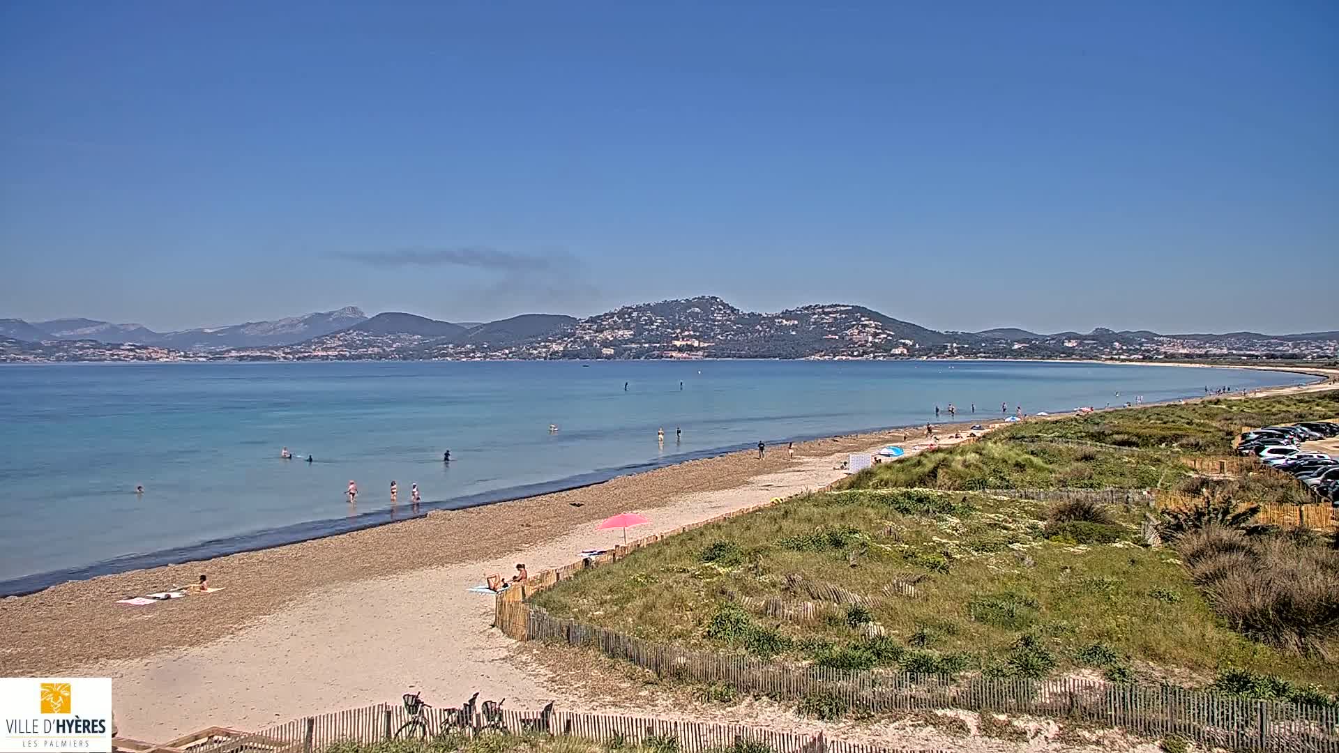 A sandy beach with a few people swimming and sunbathing stretches along a calm, light-blue sea under a clear blue sky, backed by distant mountains.