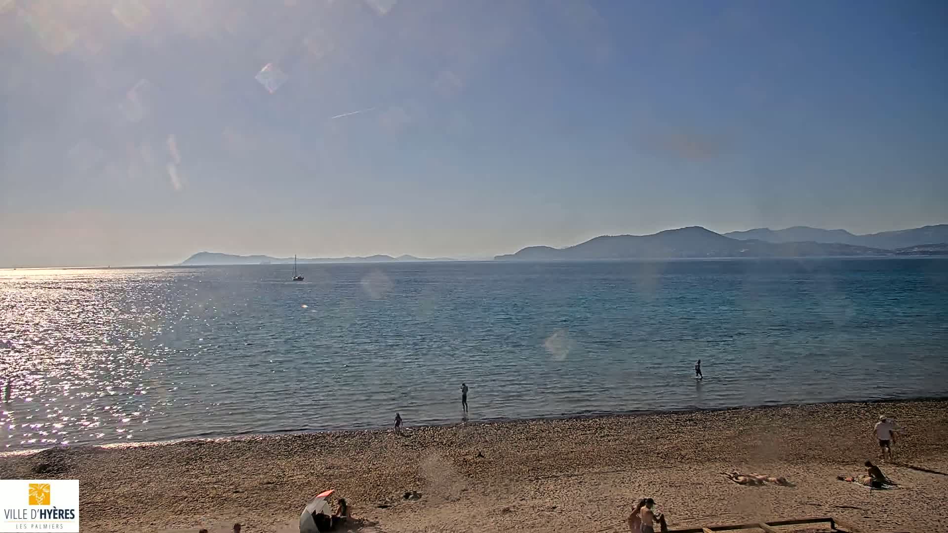 A sunny day at a pebble beach shows a calm sea with distant mountains under a mostly clear sky, a few people are visible on the beach and in the water.