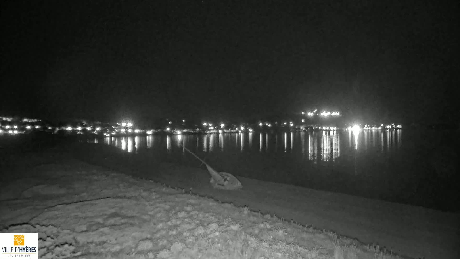 A capsized sailboat lies on a dark shoreline at night, across from a town illuminated by distant lights reflected in calm water.