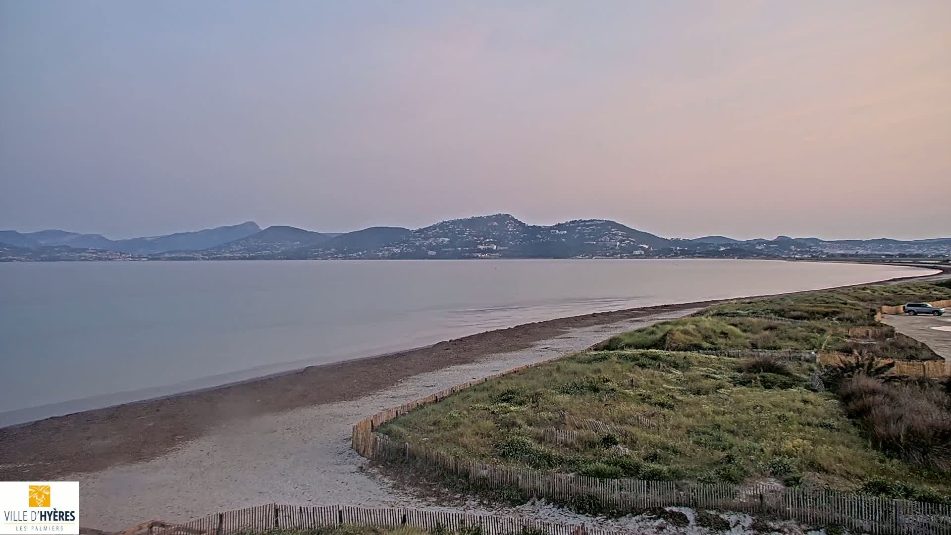 A calm, pale-lavender-hued sky overlooks a sandy beach curving gently around a body of water, with low-lying vegetation and distant mountains.