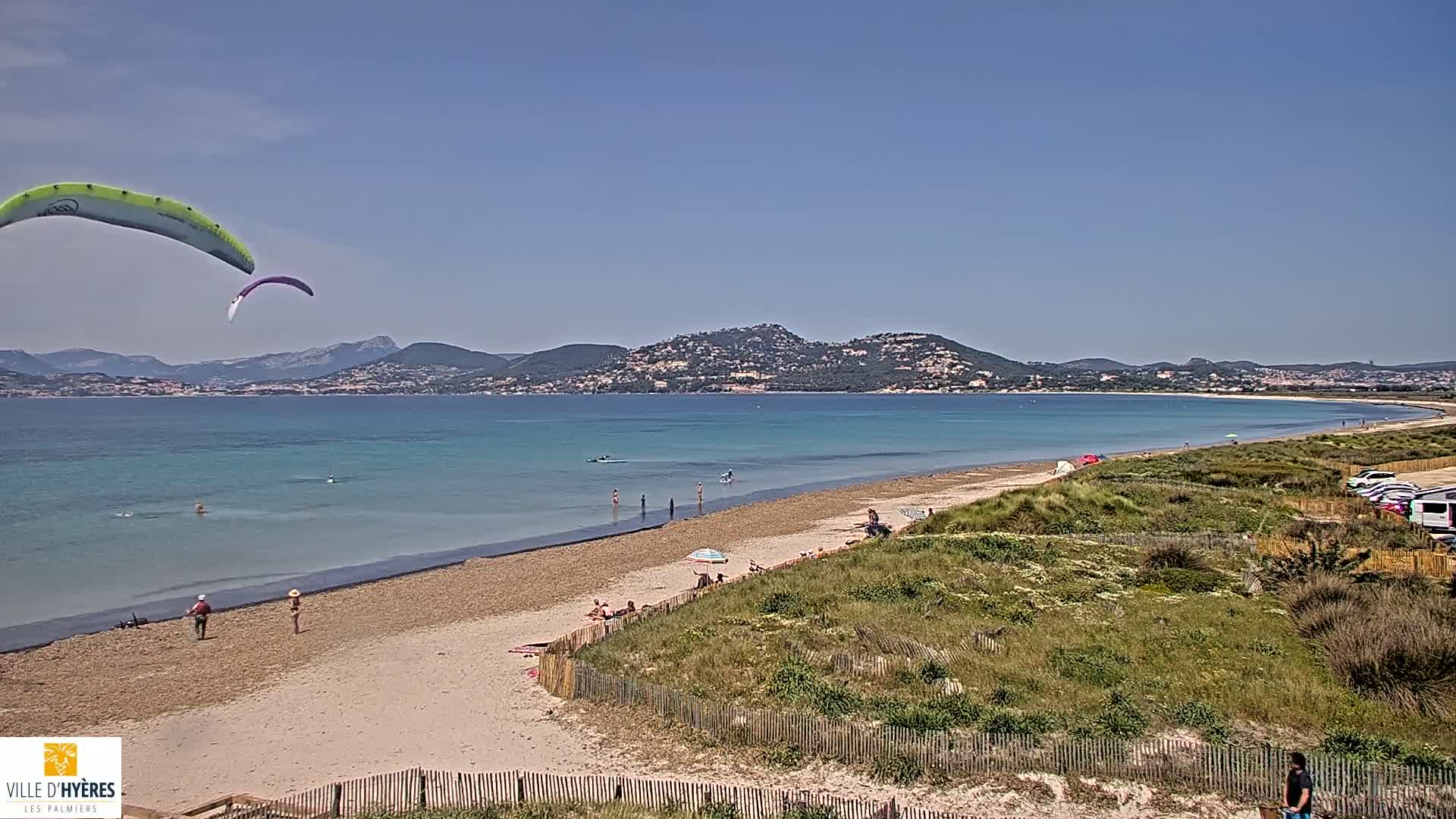 A sandy beach with a few people on it stretches along a calm, turquoise sea under a clear blue sky, with two paragliders visible in the air and mountains in the distance.