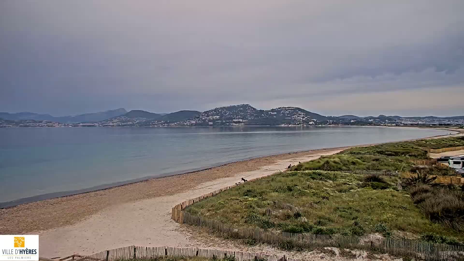 A sandy beach stretches along a calm, light-blue sea under an overcast sky, with distant hills and a town visible in the background.