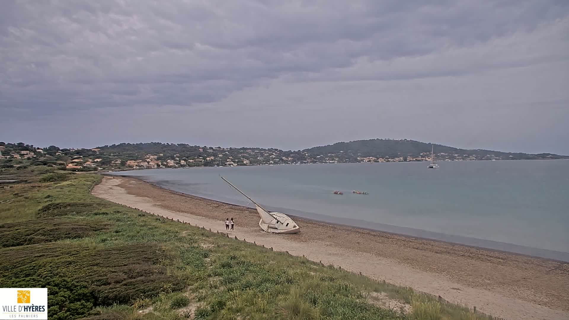 A beached sailboat rests on a sandy beach under an overcast sky, with calm water and a distant shoreline visible.