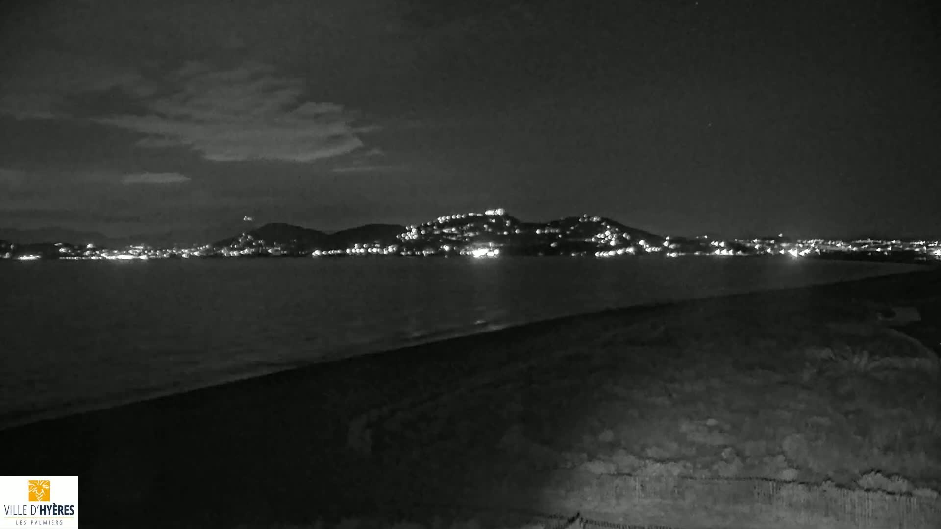 A nighttime view shows a dark beach and calm water with a city's lights illuminating hills in the distance under a cloudy sky.
