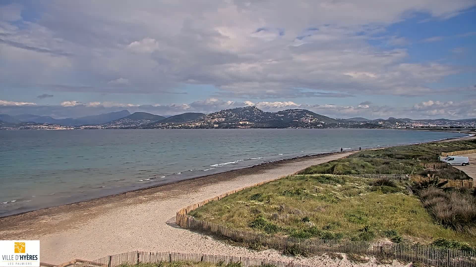 A partly cloudy day shows a sandy beach with a wooden fence separating it from grassy dunes, overlooking a calm bay and distant mountains.