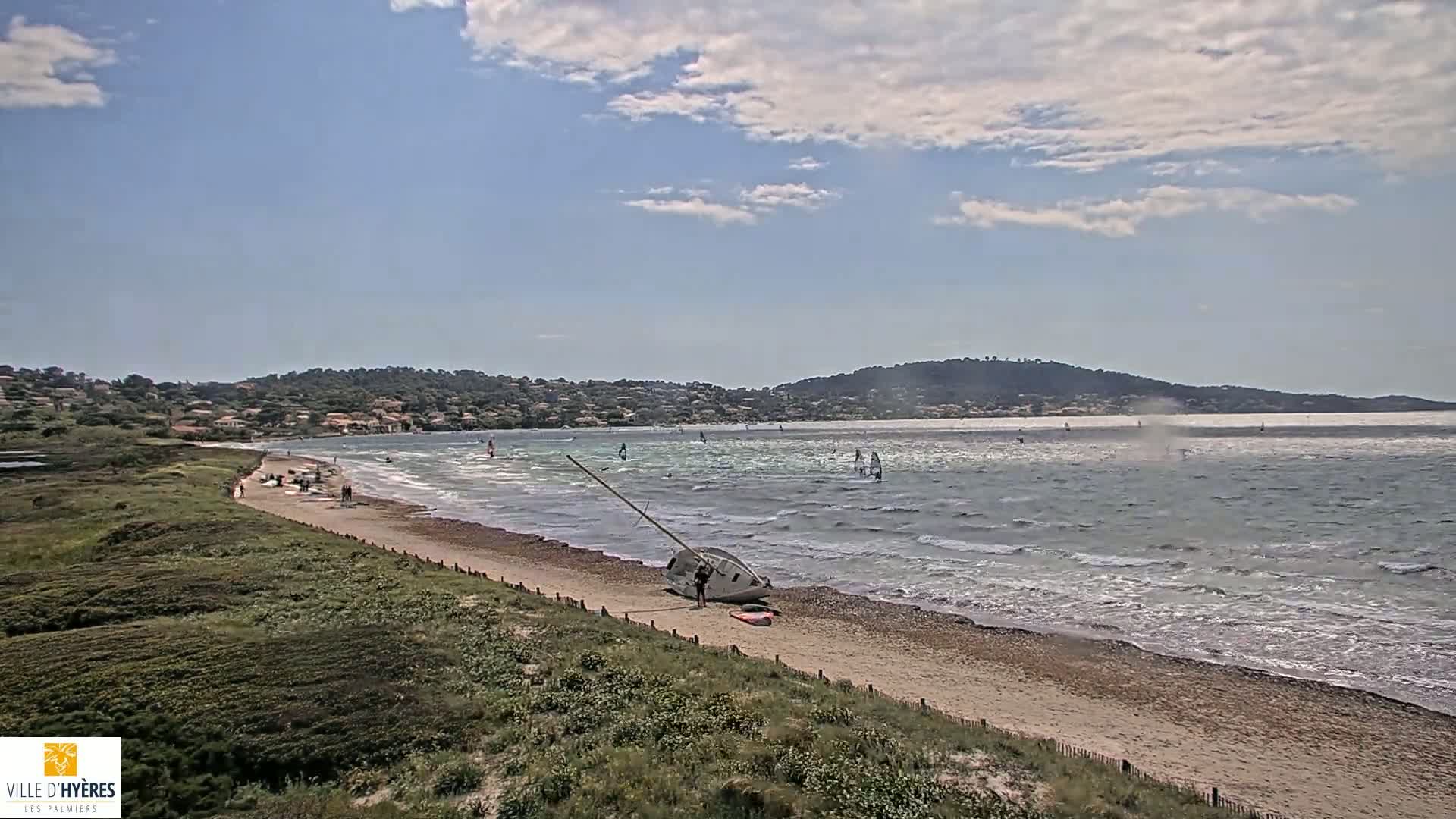 A sandy beach with a capsized sailboat and several windsurfers on a partly cloudy day.