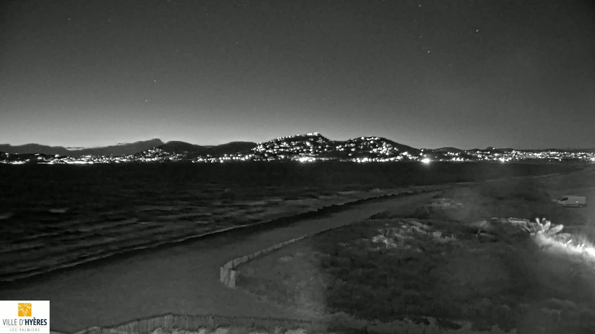 A nighttime, black and white view of a wavy ocean shore with a hilltop town illuminated in the distance under a starry sky.