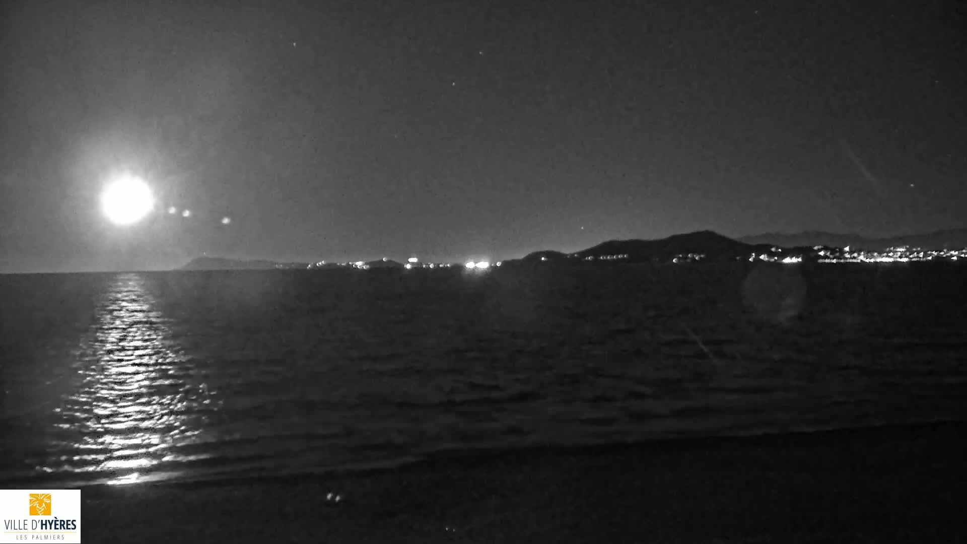 A bright moon illuminates a calm sea at night, reflecting on the water's surface, with a distant shoreline featuring city lights visible on the horizon.