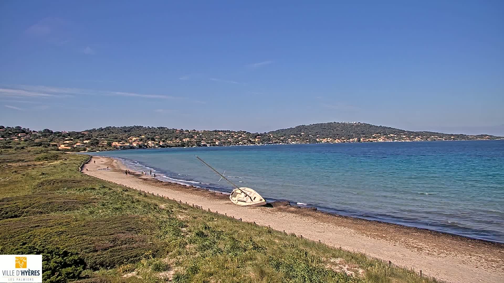 A sandy beach with a capsized sailboat rests on the shore, overlooking a calm, turquoise sea and a distant hilly coastline under a clear blue sky.