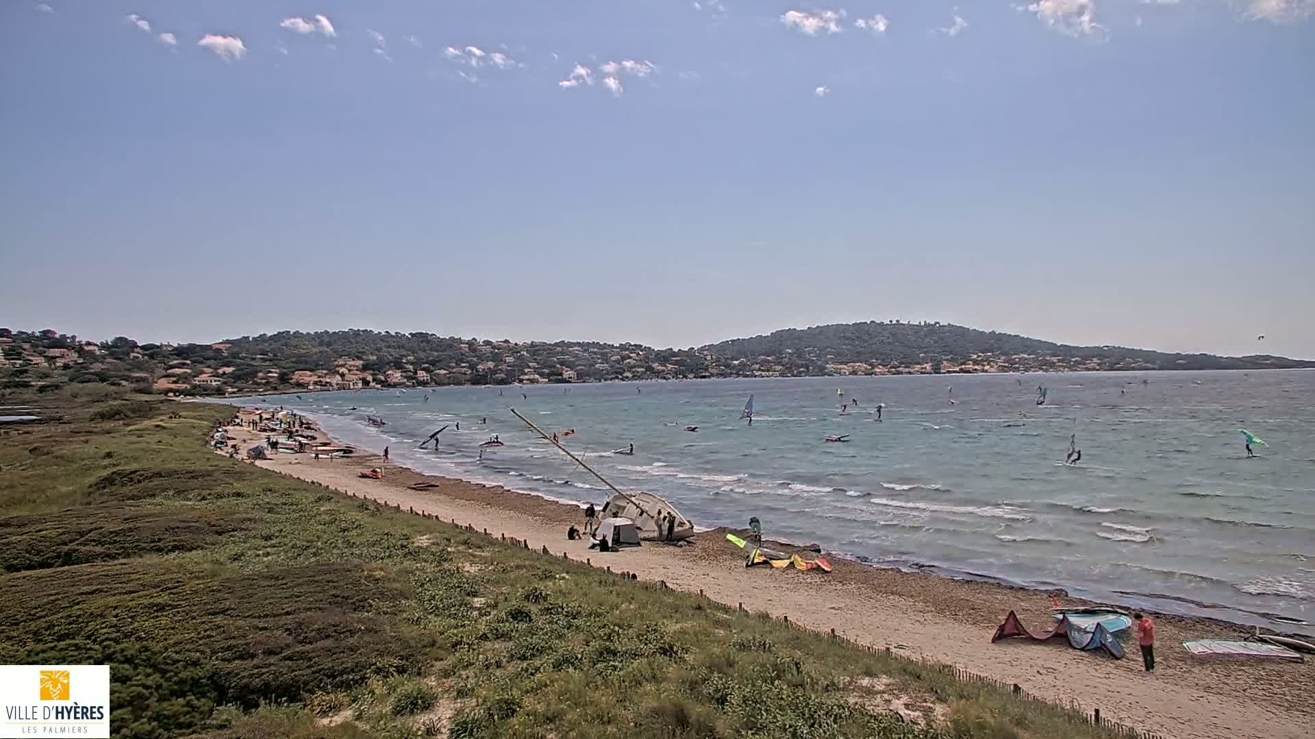 A sandy beach with people windsurfing and kitesurfing in a calm sea under a partly cloudy sky, backed by a low hill and distant houses.