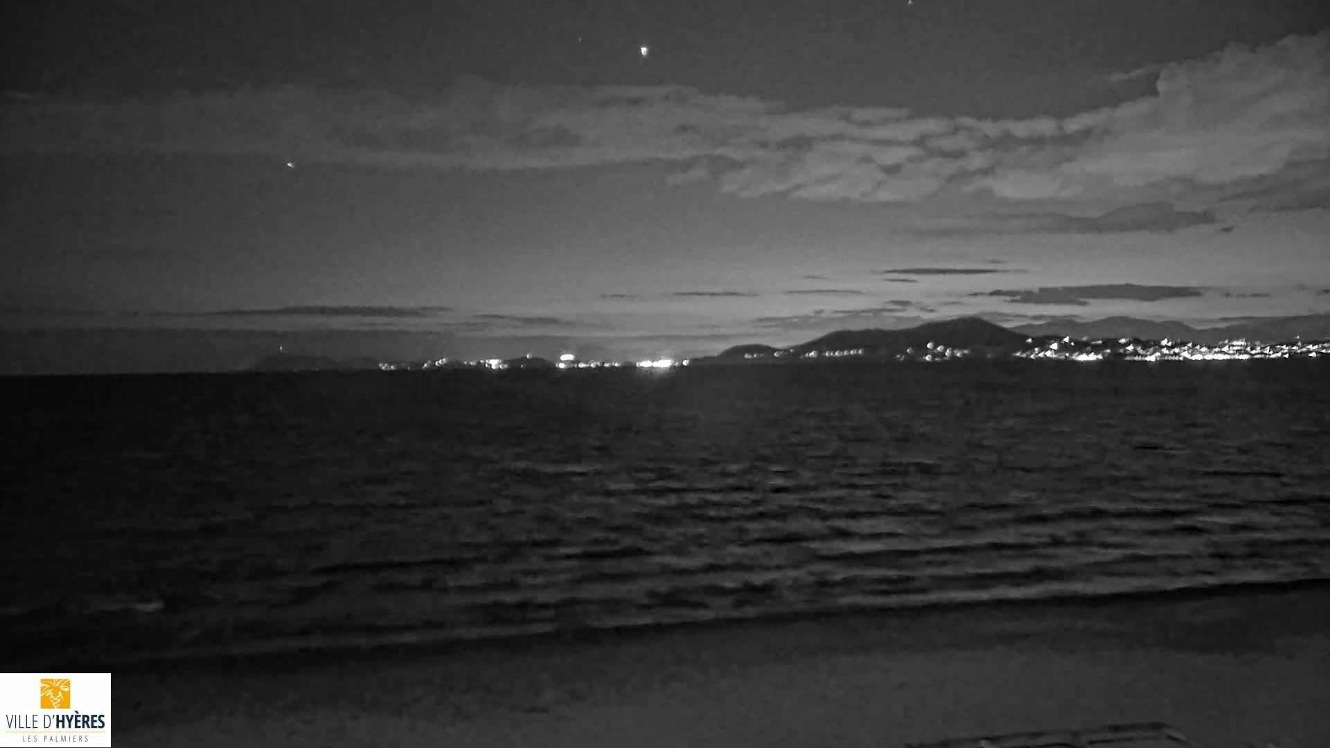 A nighttime, black and white view of a calm sea with a distant, illuminated city skyline under a cloudy sky.