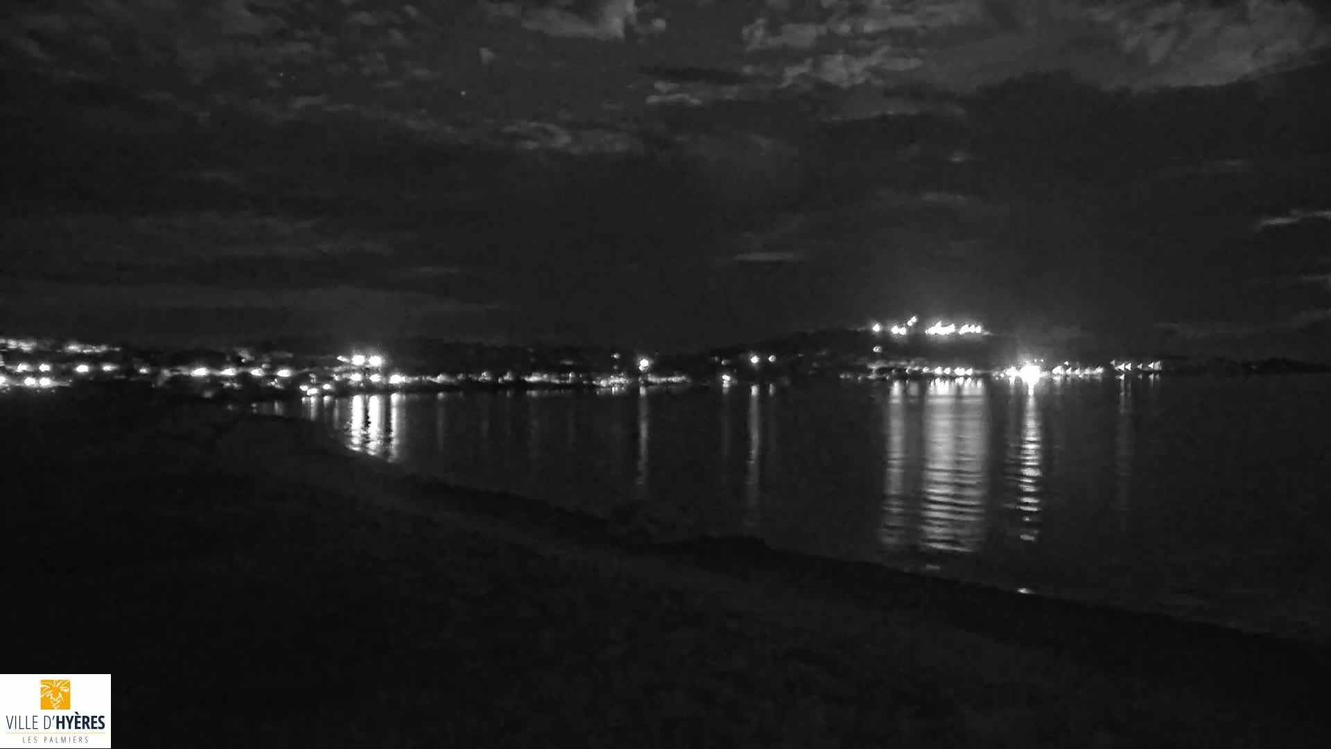 A nighttime view of a calm body of water reflecting the lights of a distant town under a cloudy sky.