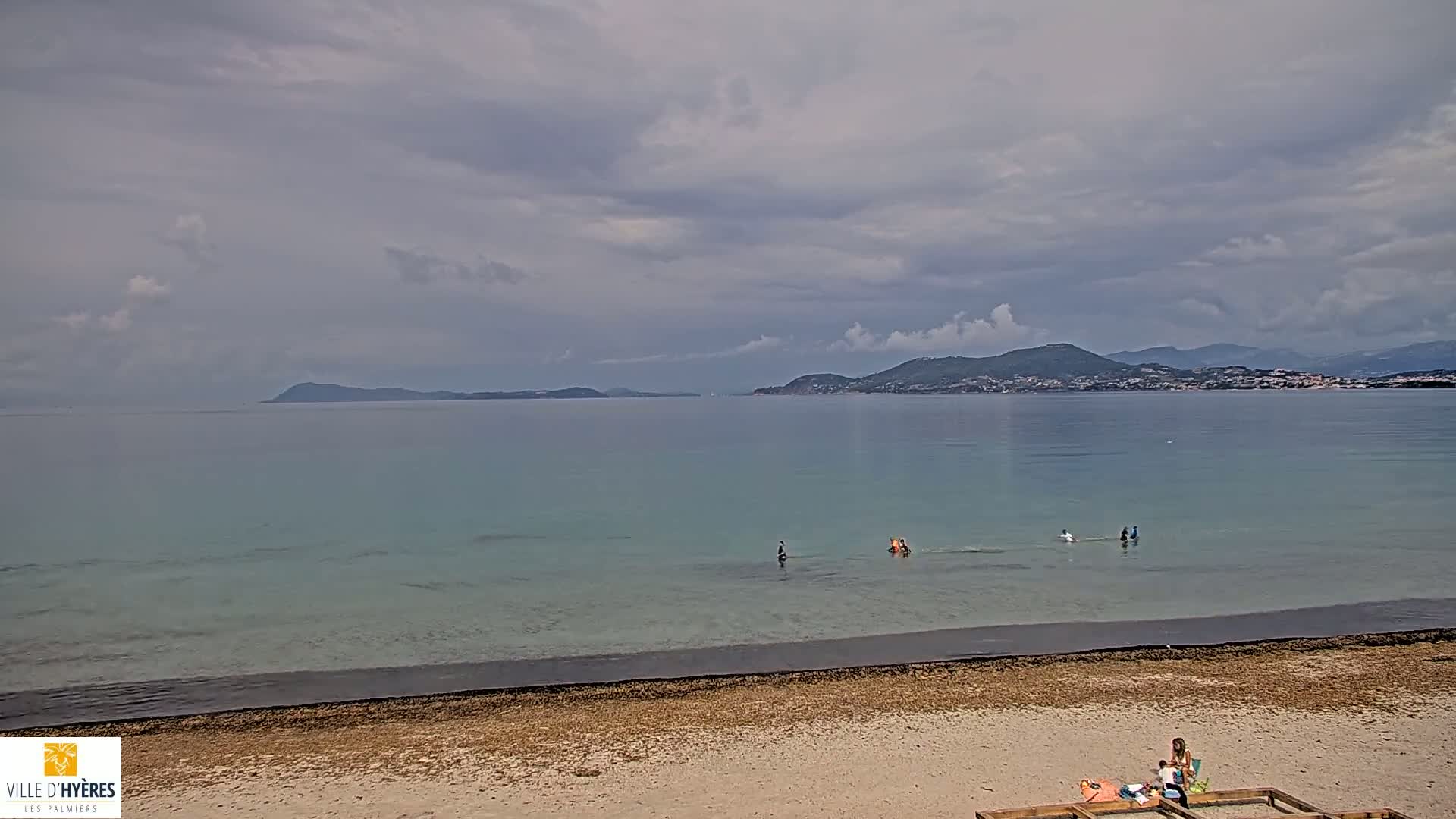 A sandy beach with a few people wading in shallow, clear water, overlooks a calm sea and distant mountains under a cloudy sky.