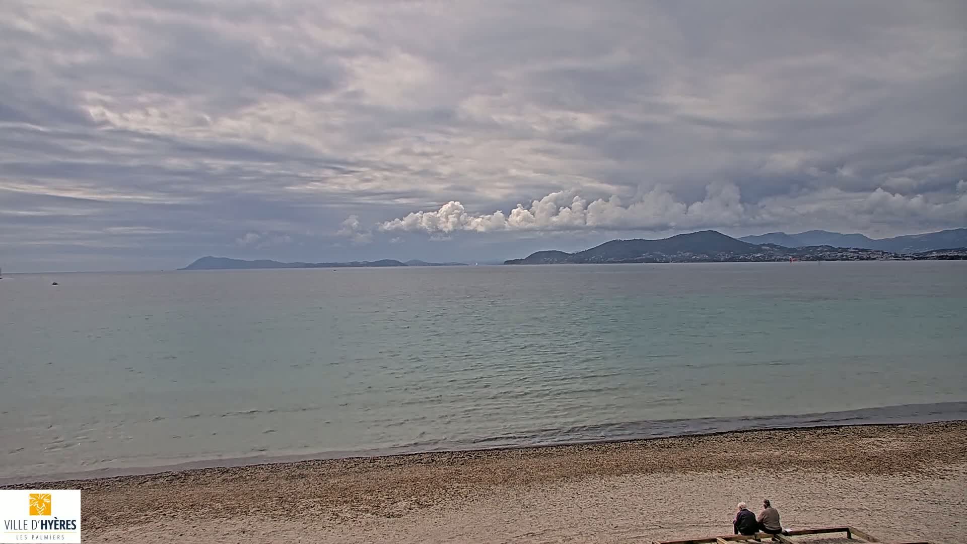 A calm, pale-green sea stretches to distant mountains under a mostly cloudy sky, with two people seated on a bench at a pebble beach in the foreground.