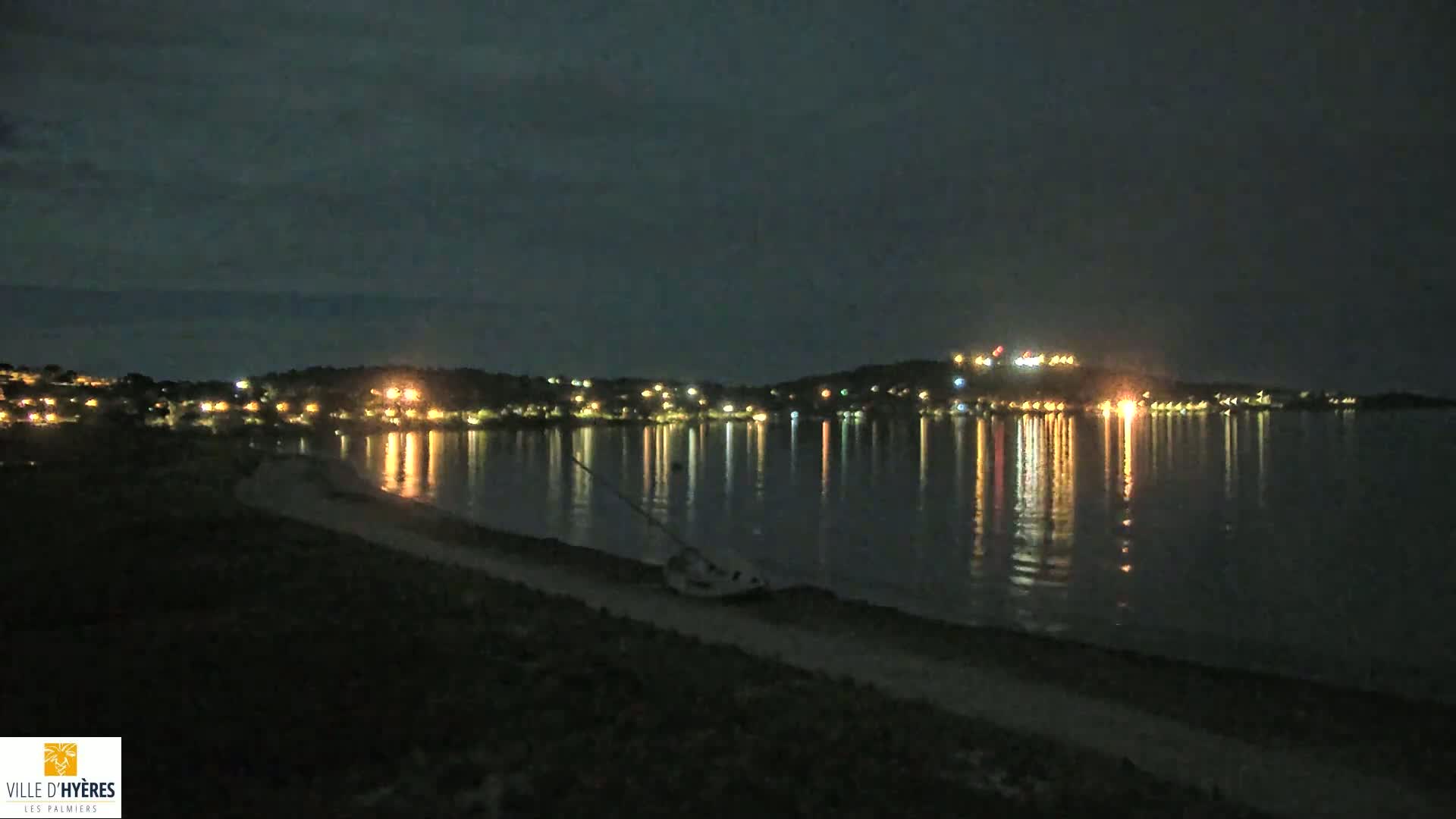 At night, a dark shoreline with a beached sailboat is visible, looking across a calm body of water reflecting the lights of a distant town on a hill.