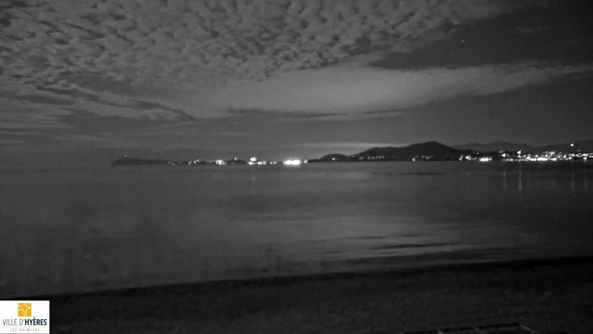 A dark, nighttime beach scene shows a calm body of water reflecting the lights of a distant city under a cloudy sky.