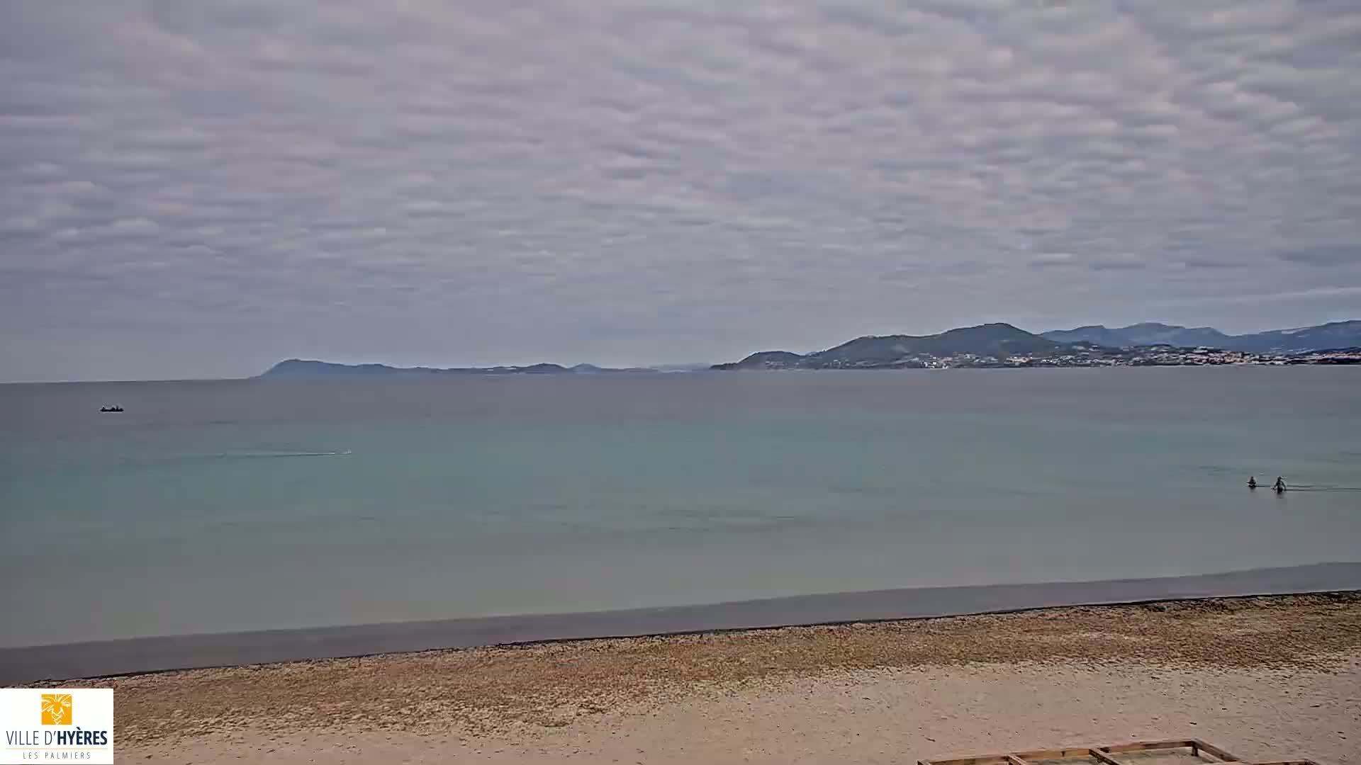 A sandy beach with calm, light-green water stretches to a distant shoreline under a cloudy sky, where two people are wading in the water.