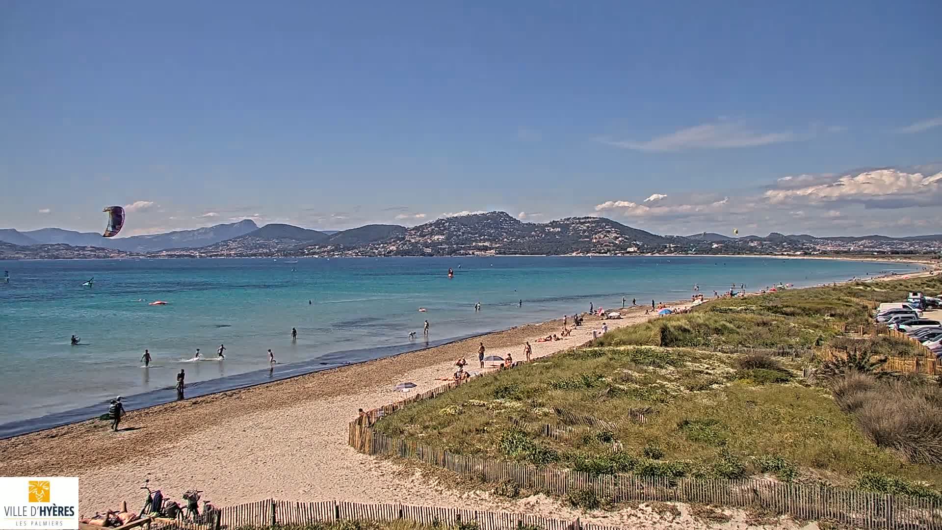 A sandy beach with people swimming and sunbathing, a kite surfer in the water, and a mountainous landscape in the background under a mostly sunny sky.