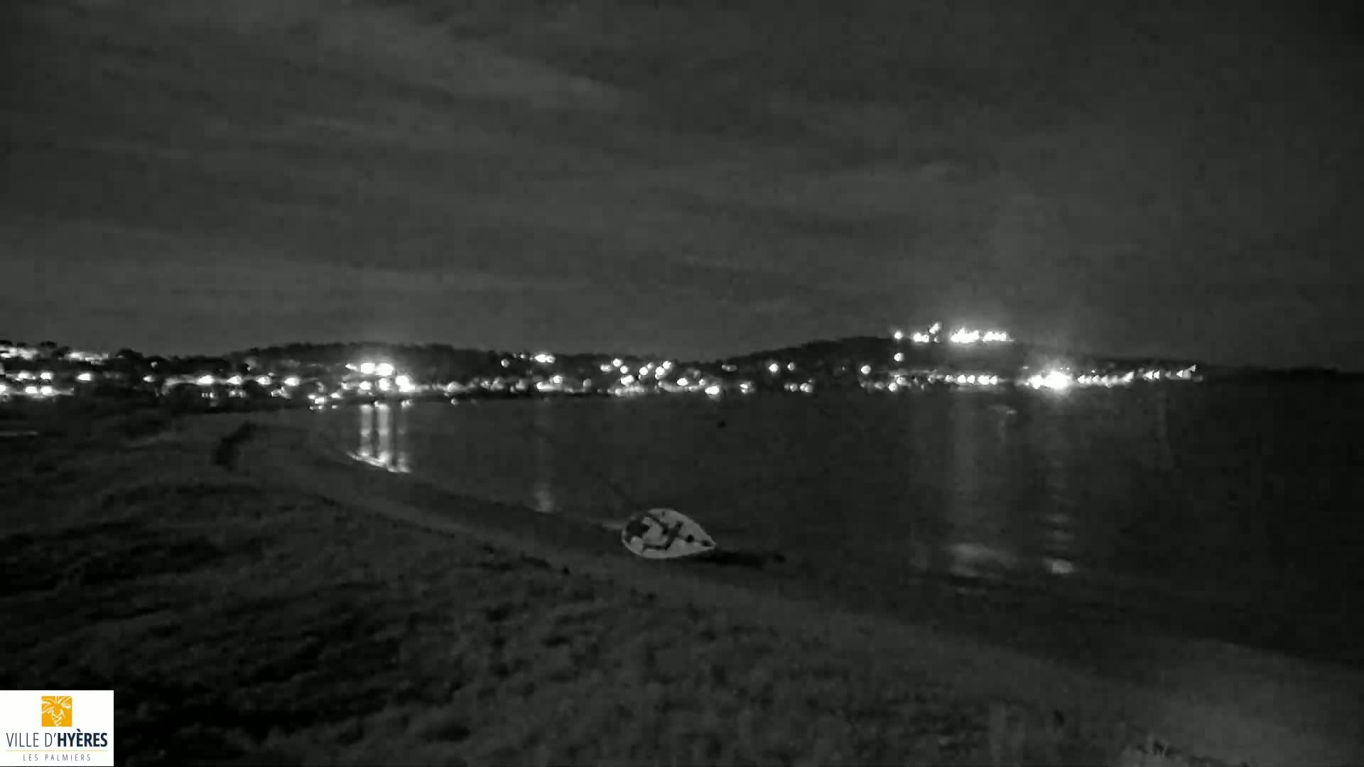 A small boat lies on a dark beach at night, under a cloudy sky, across from a dimly lit town.