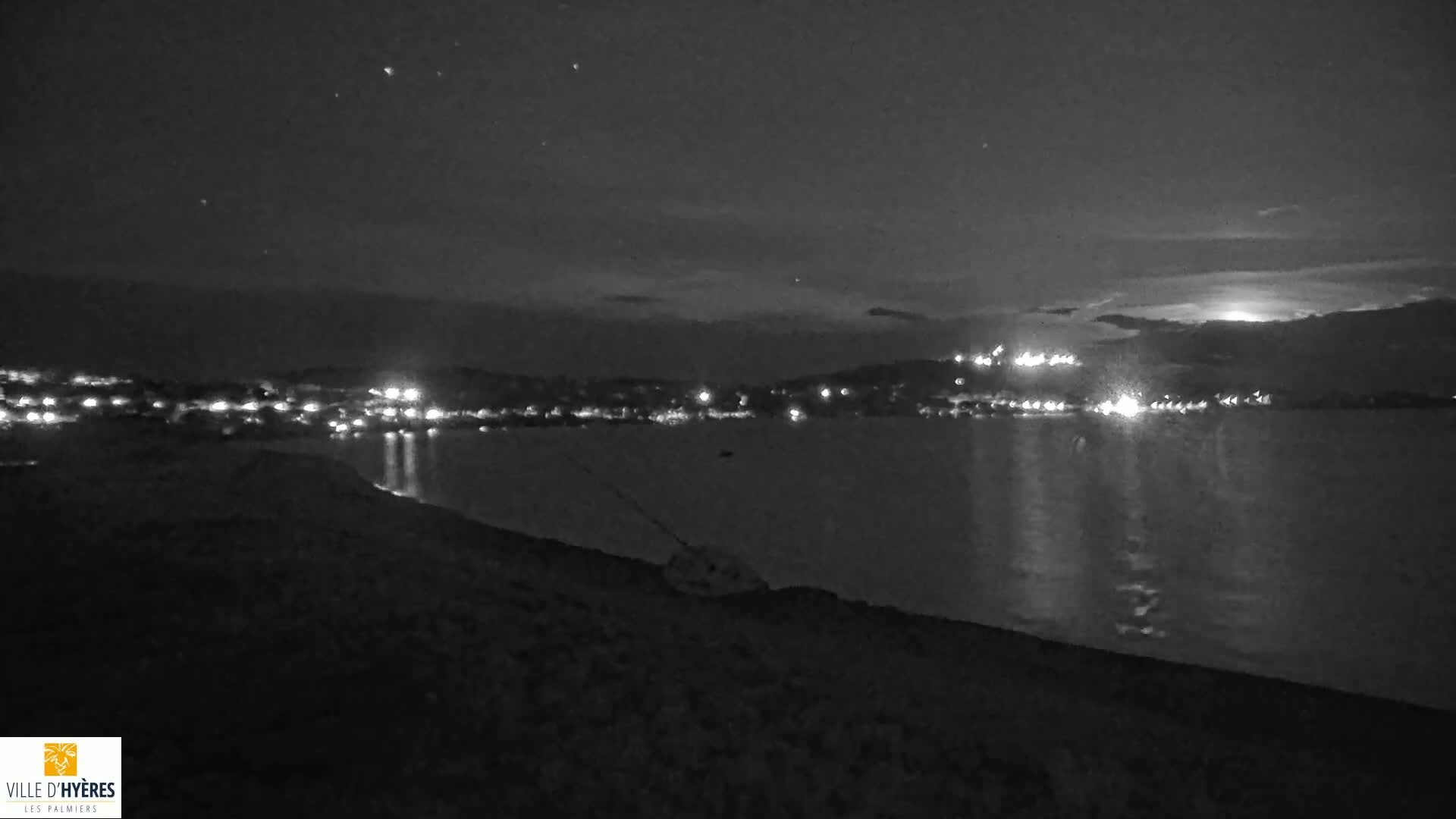 A nighttime view of a calm body of water reflecting the lights of a distant town under a clear, starry sky.