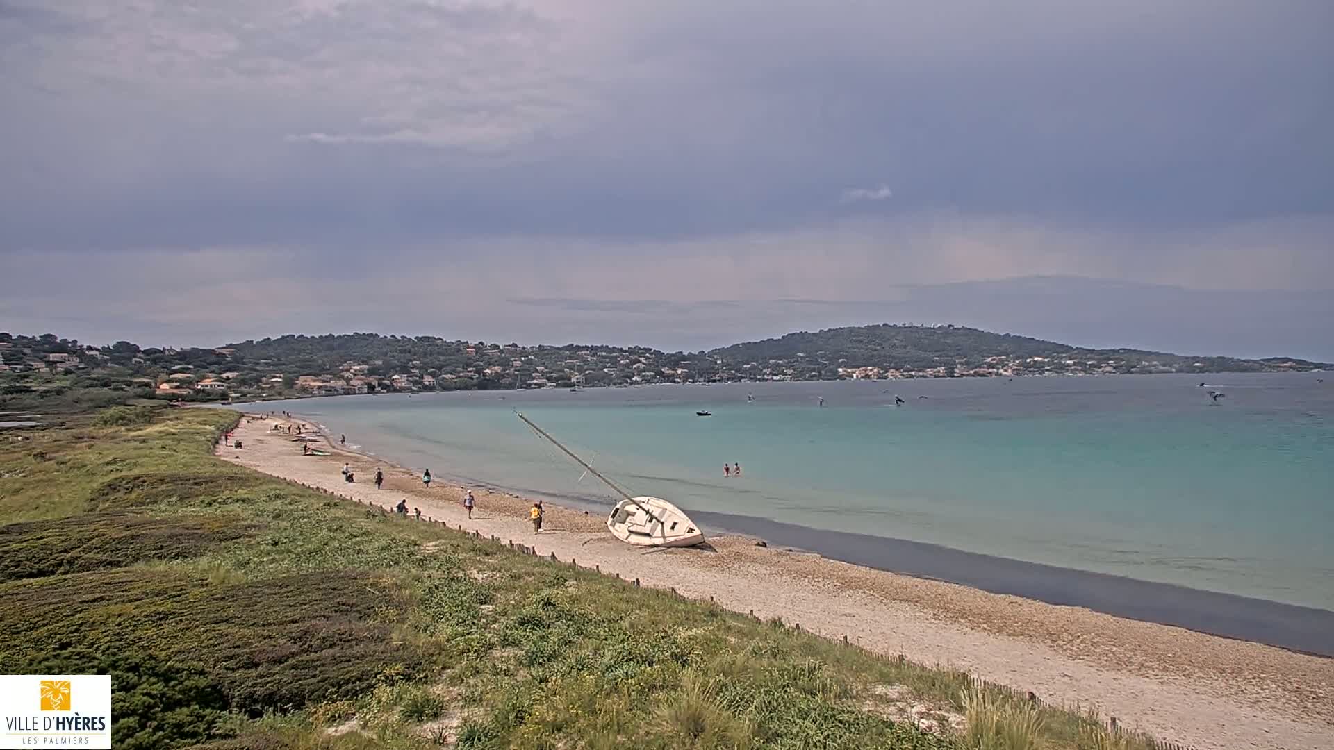 A sandy beach with a beached sailboat and several people walking along the shore, under an overcast sky, overlooks a calm, turquoise bay with distant hills and a few boats.