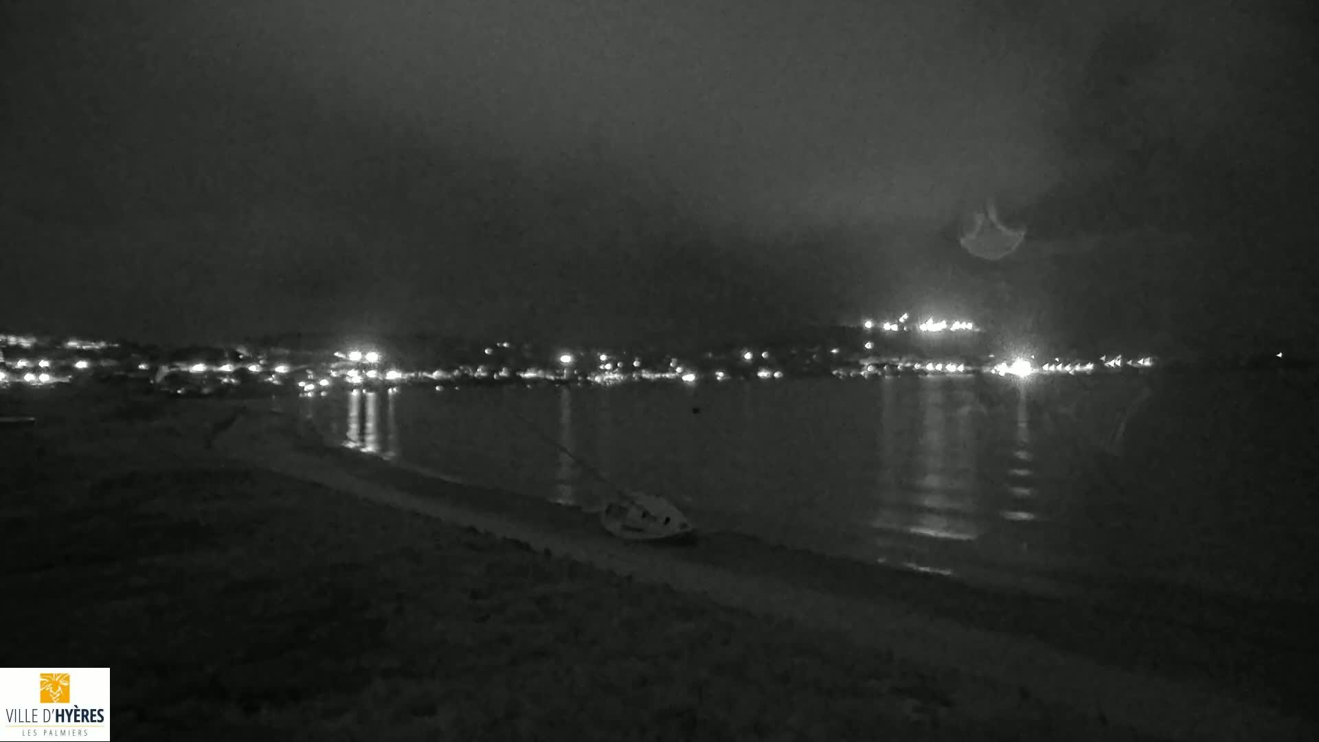 A dark, nighttime beach scene shows a distant town illuminated by lights reflected on calm water, with a small boat beached on the shore.
