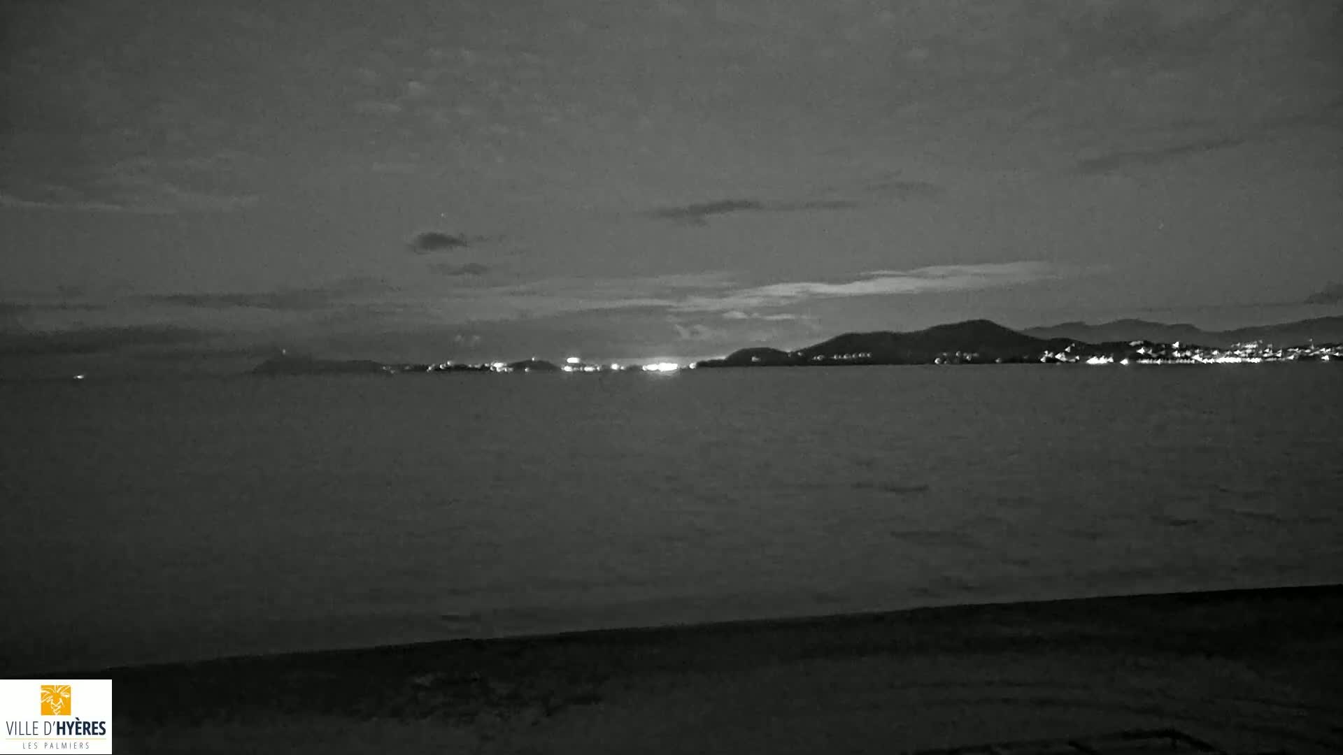 A nighttime, grayscale view shows a calm body of water with a distant, dimly lit shoreline under a cloudy sky.