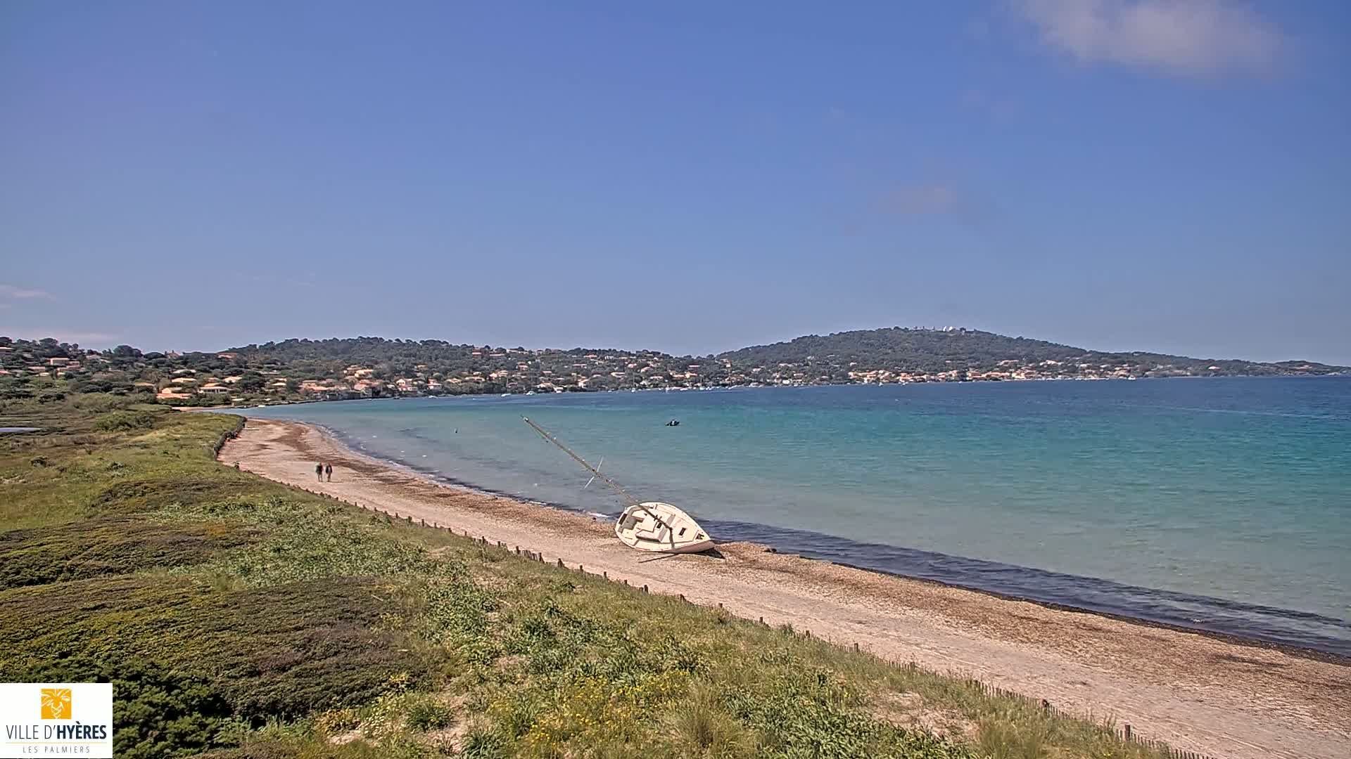 A sandy beach with a capsized sailboat lies on the shore, next to a calm, turquoise sea under a clear blue sky.