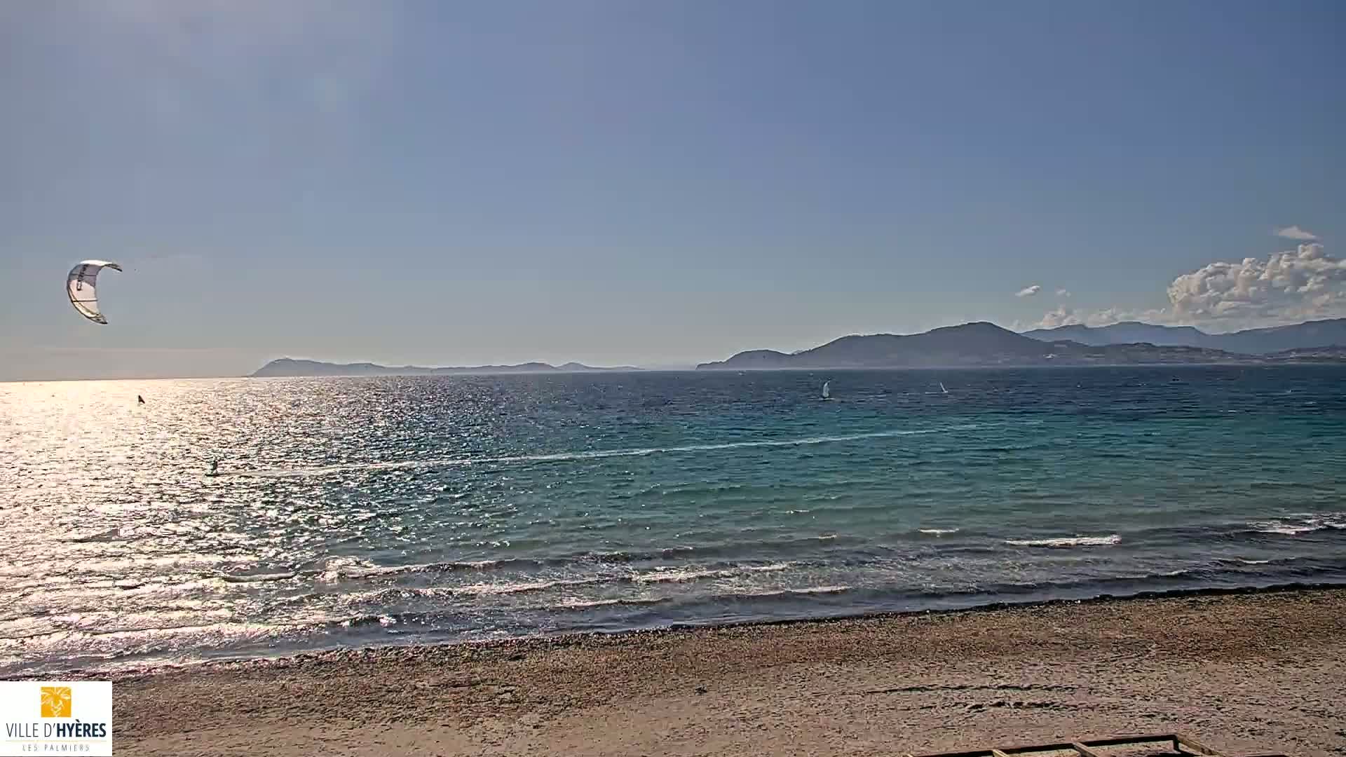 A sandy beach overlooks a calm, turquoise sea with a kiteboarder and sailboats visible in the distance under a mostly sunny sky.