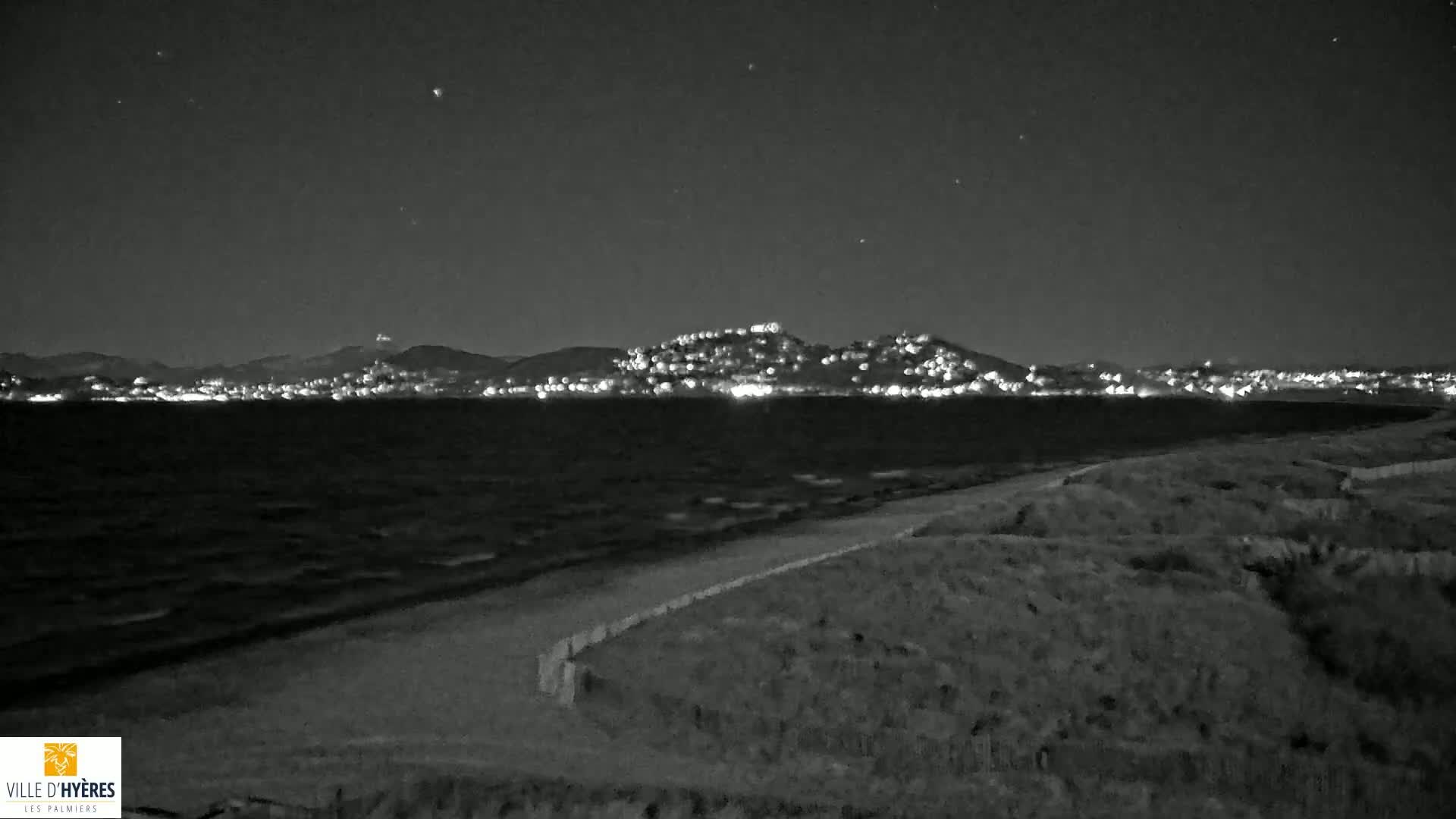 A moonlit, nighttime beach scene shows a dark, wavy ocean, a sandy shore, and a distant city glowing with lights against a mountainous backdrop.