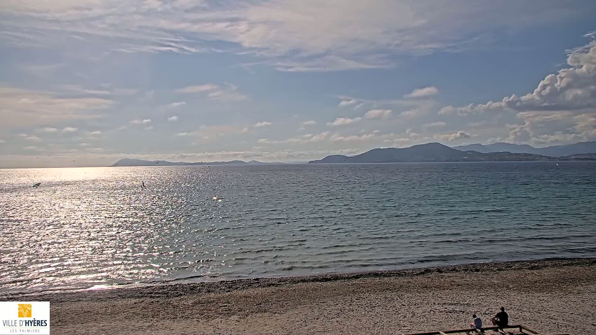 A sandy beach overlooks a calm, sparkling sea with distant mountains under a partly cloudy sky.