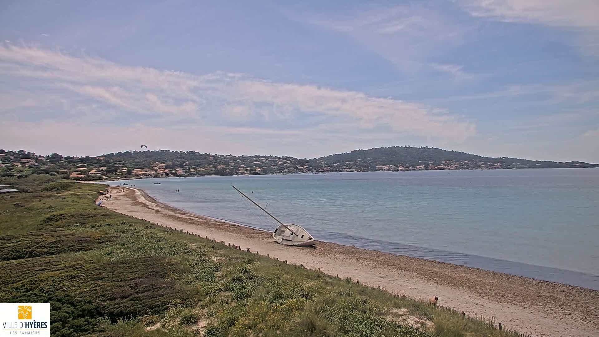 A partly cloudy day shows a sandy beach with a capsized sailboat, a few people, and a calm, light-blue sea with a distant coastline.