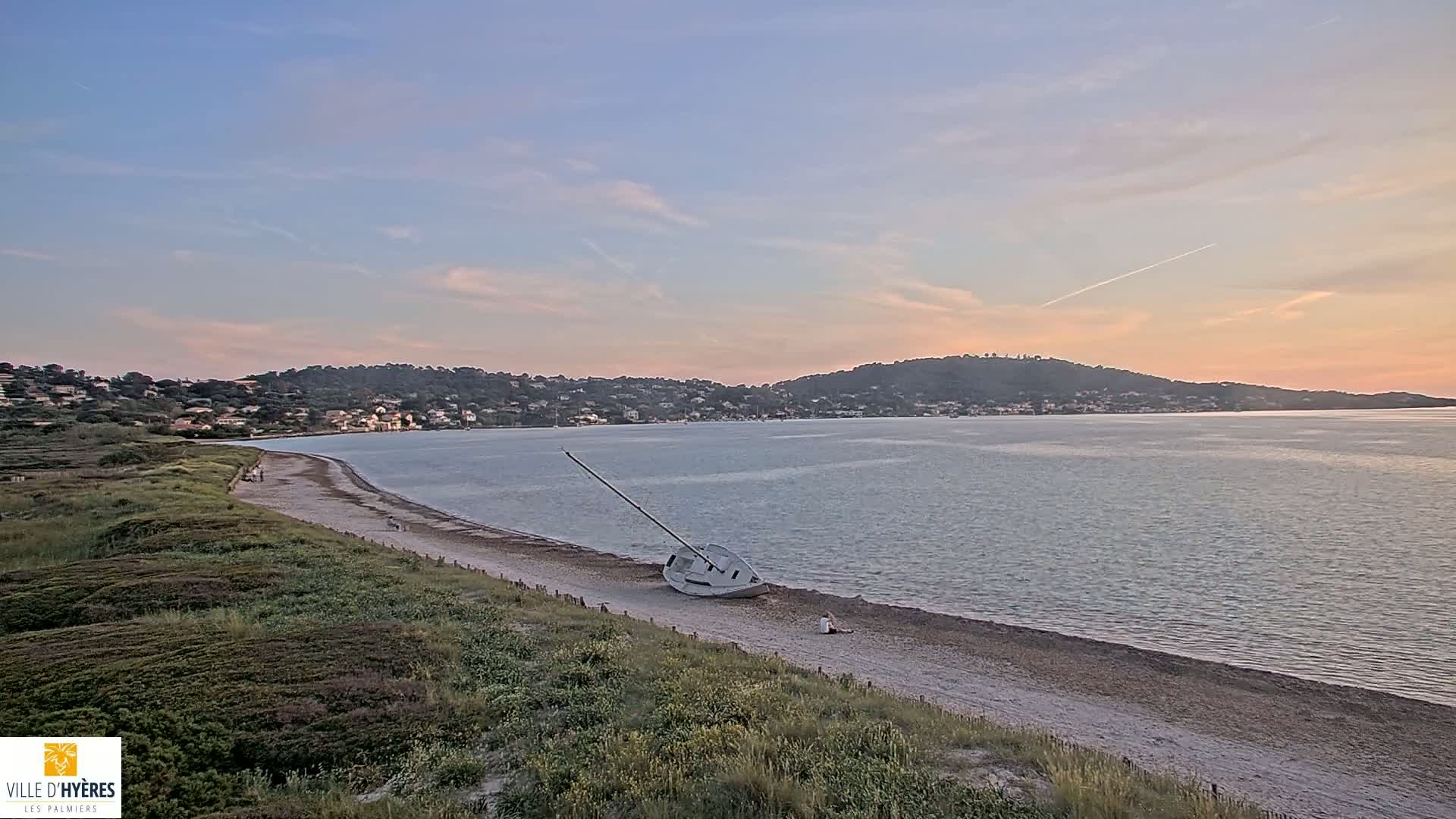 A capsized sailboat lies on a sandy beach under a pale sunset sky, with a calm body of water and distant hills in the background.