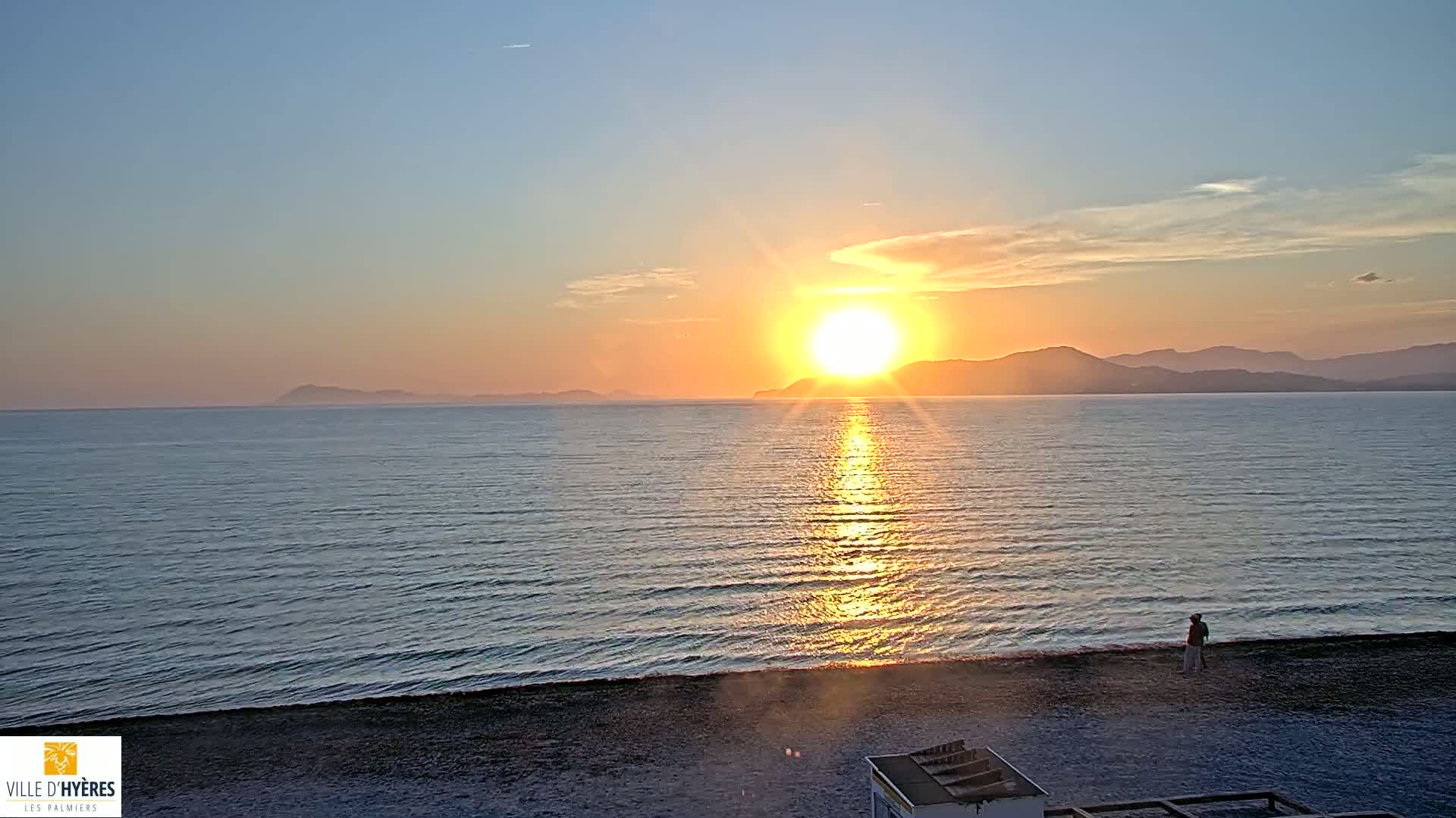 A person stands on a dark-sand beach watching a bright orange sunset over a calm sea and distant mountains.