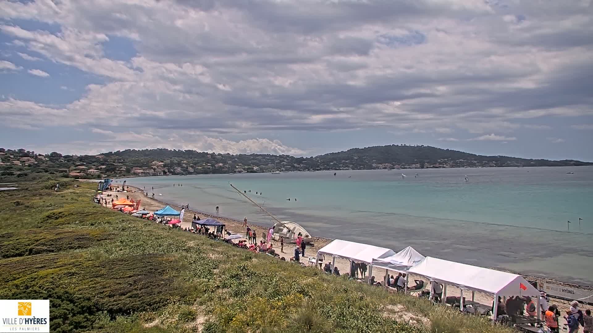 A sandy beach with people, tents, and a capsized sailboat, under a partly cloudy sky, overlooks a calm, turquoise bay and distant hills.