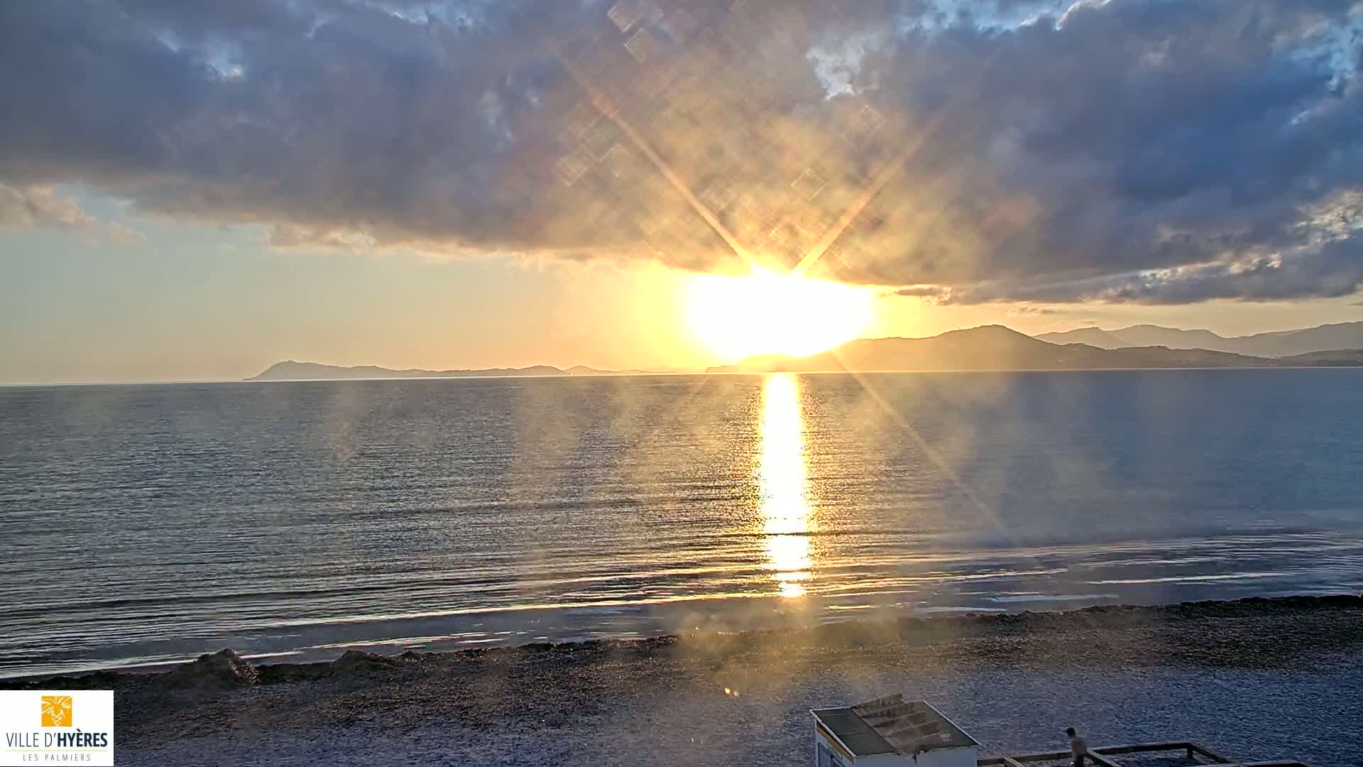 A bright sun shines through partly cloudy skies over a calm sea and a pebble beach at sunset.