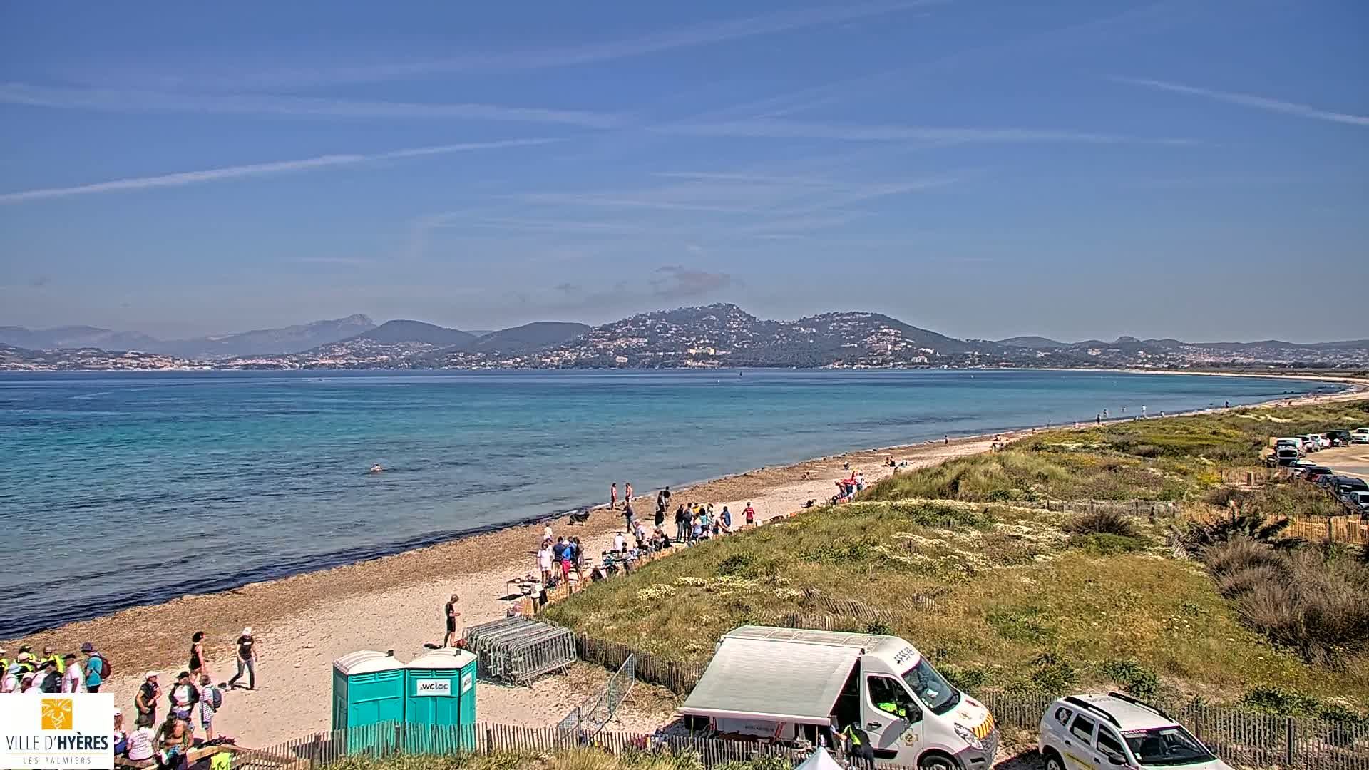 A sandy beach with a few people on it stretches along a calm, turquoise ocean under a clear blue sky, with distant mountains visible across the water.