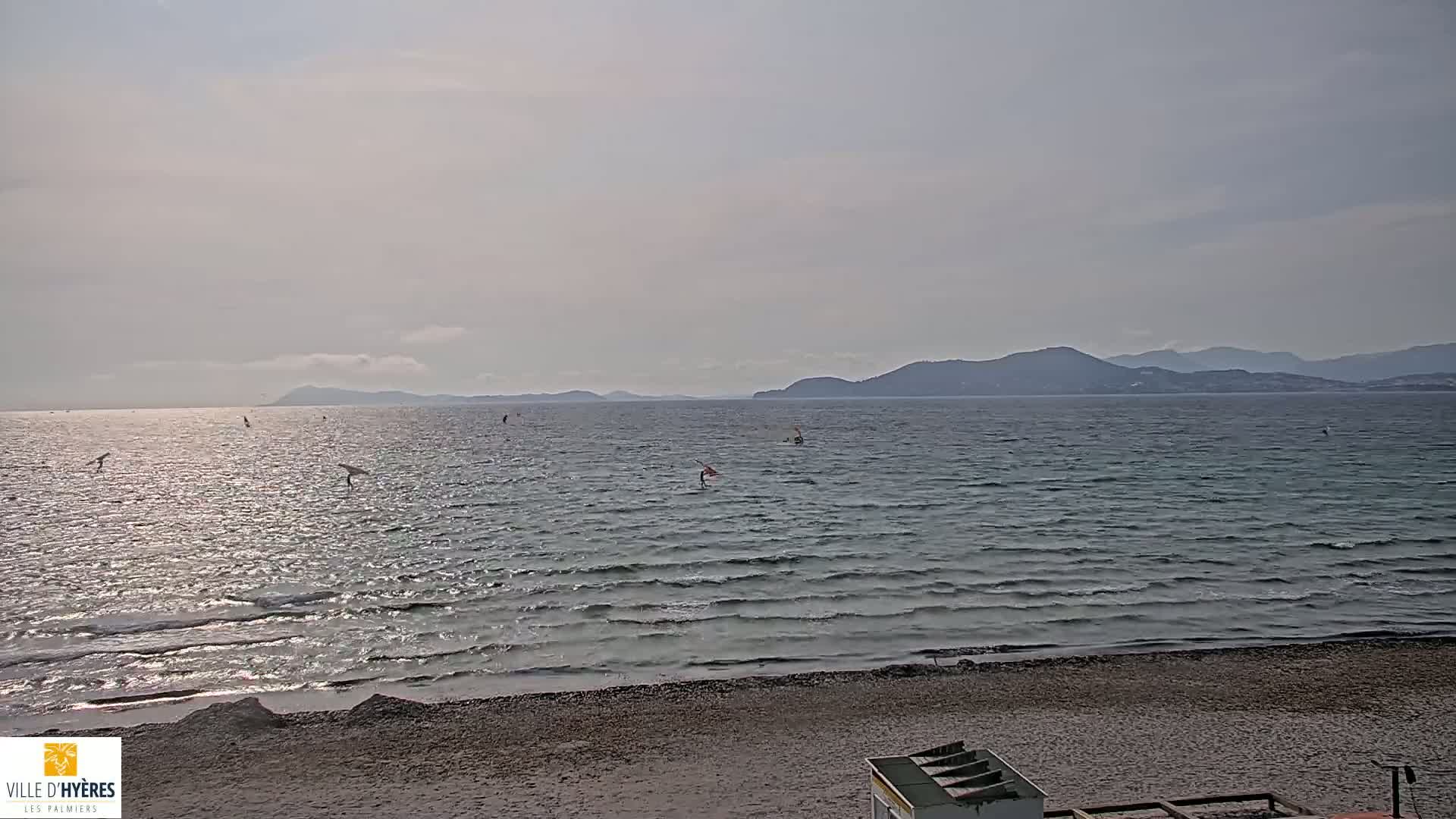 A sandy beach with several windsurfers visible on a somewhat choppy sea under a mostly cloudy sky with distant mountains.