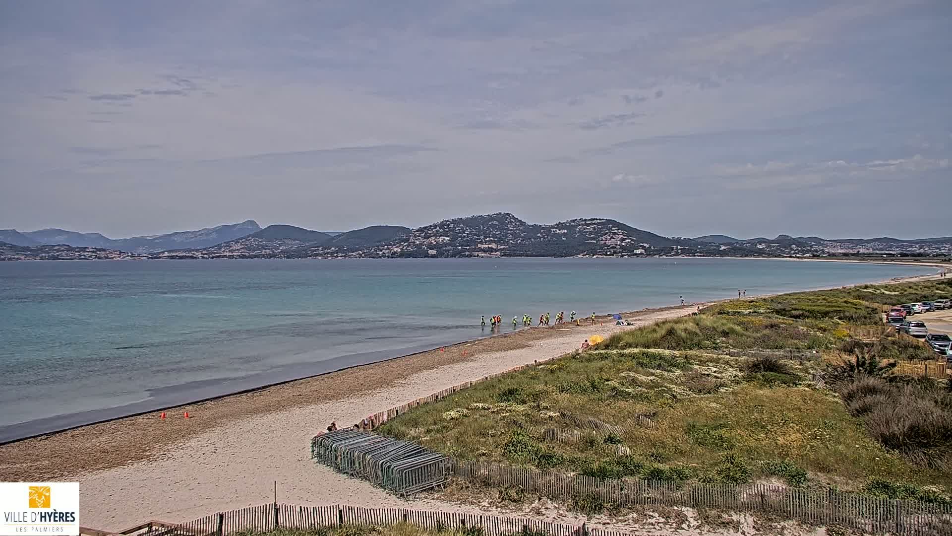 A sandy beach curves along a calm, turquoise sea, backed by a line of low hills under a mostly cloudy sky.