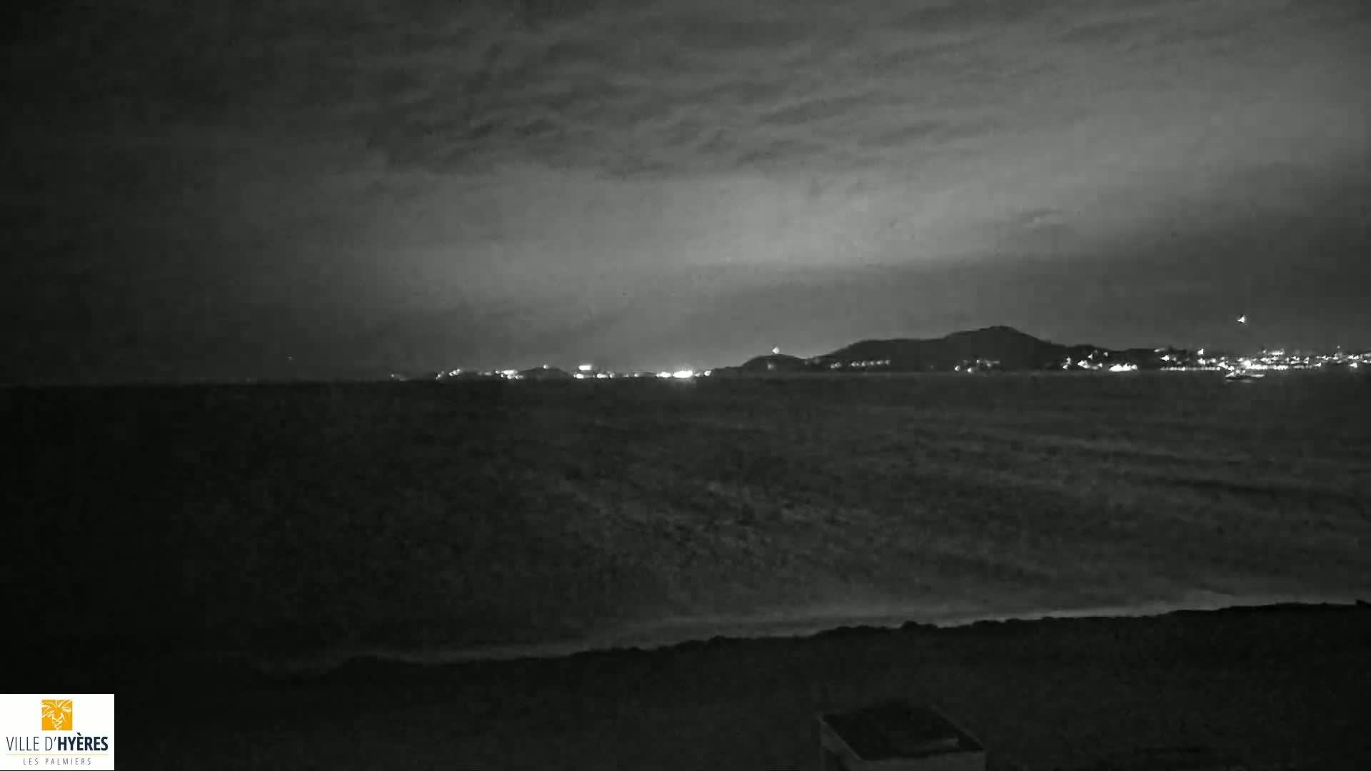 A nighttime coastal scene shows a dark, wavy sea under a cloudy sky, with a distant, dimly lit town and hills visible on the horizon.