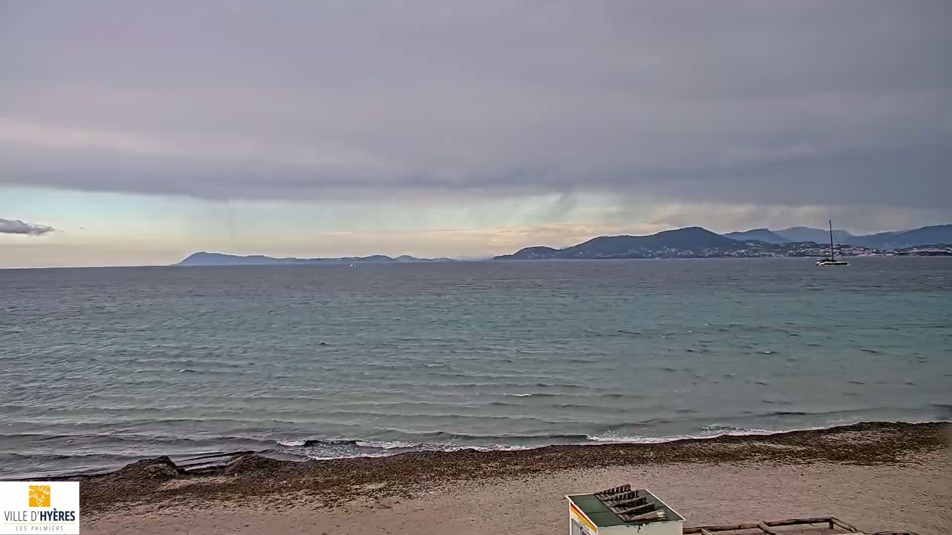 A sandy beach with small waves and a calm sea stretches towards distant mountains under an overcast sky, with rain visible in the distance.