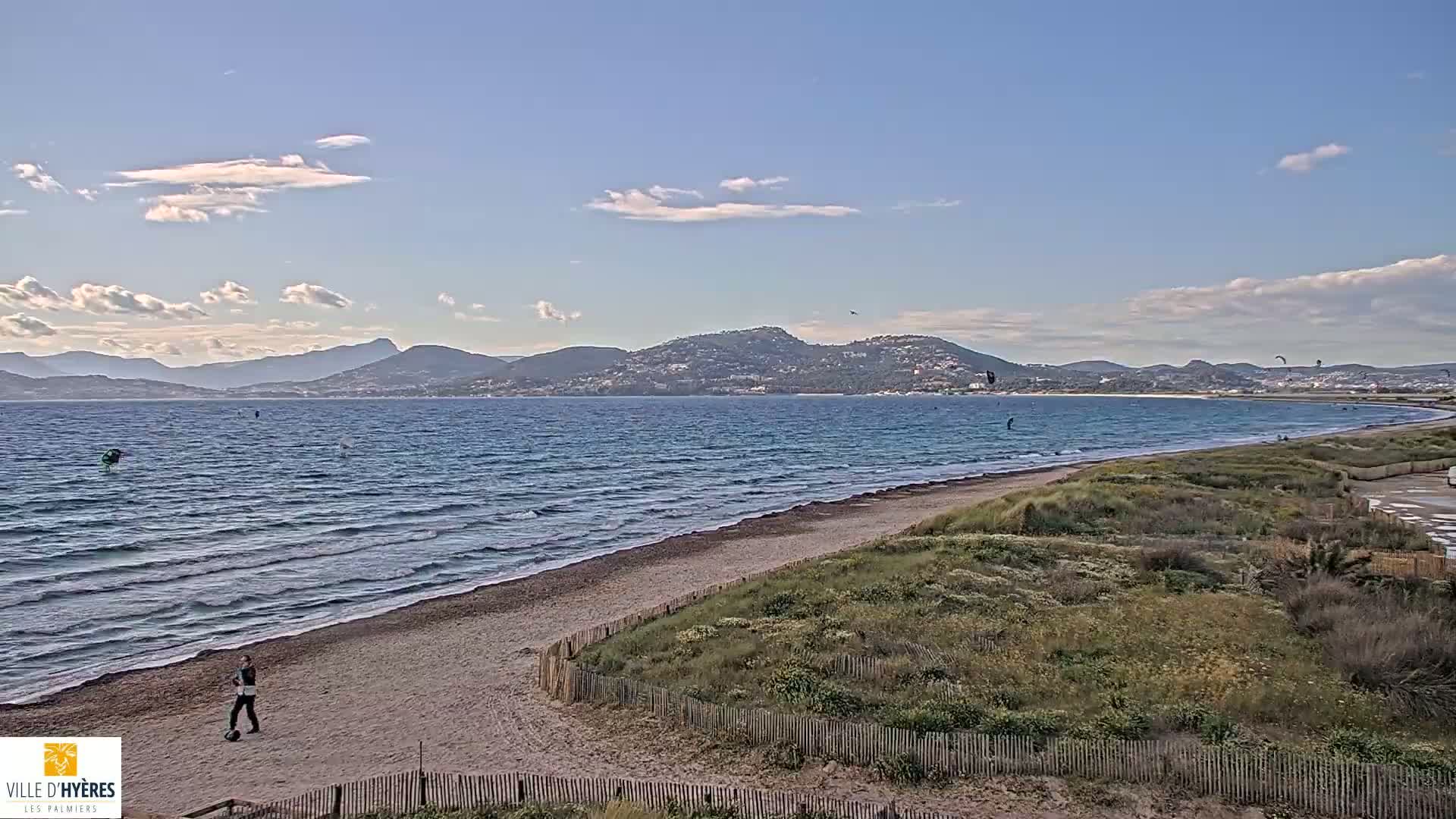 A sandy beach with gently rolling waves under a partly cloudy sky stretches towards distant mountains, with a few kitesurfers visible in the water and a person walking on the shore.