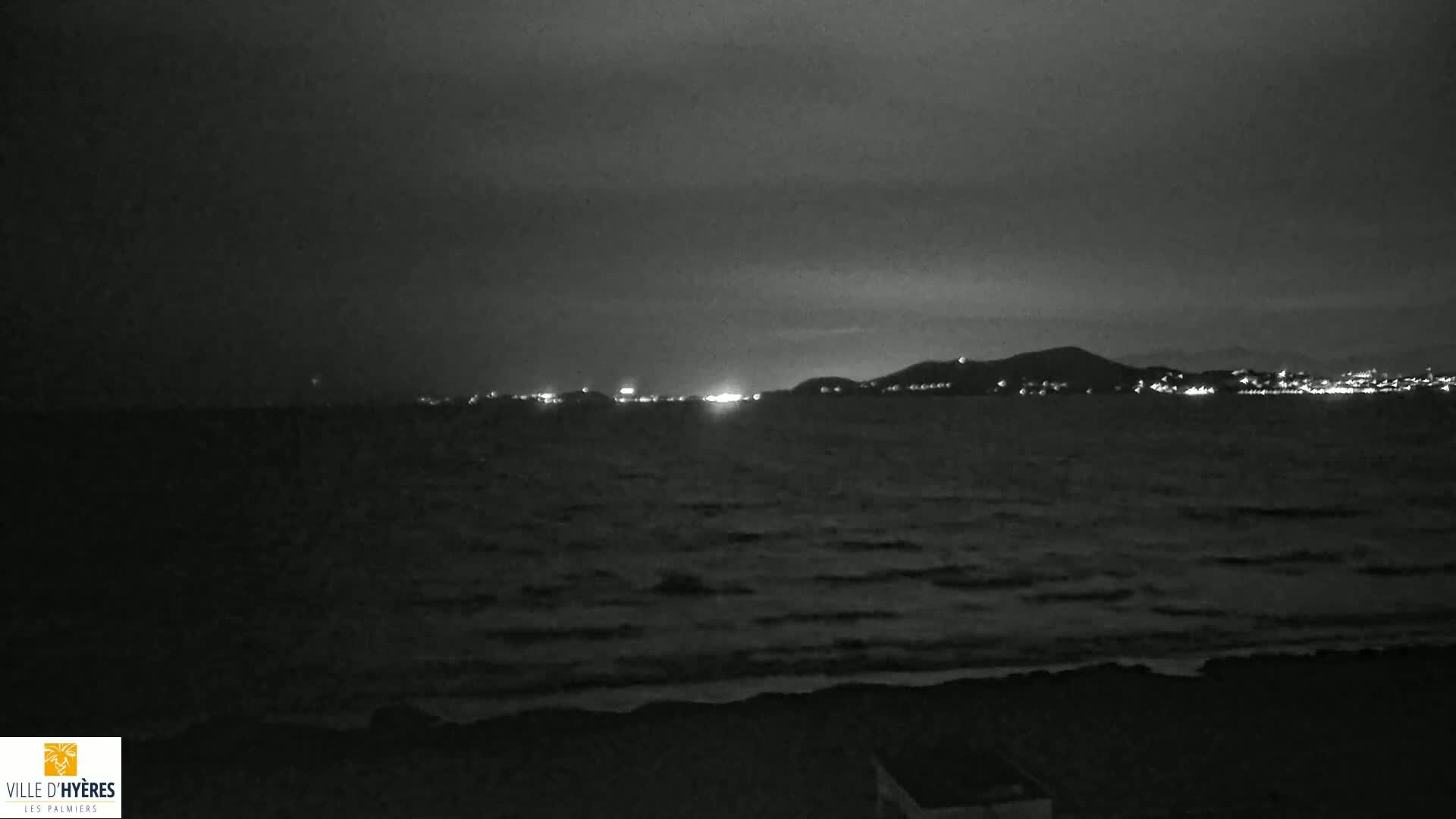 A dark, nighttime beach scene shows a calm sea with small waves lapping the shore, and a distant, faintly lit city nestled against a low mountain range under a cloudy sky.