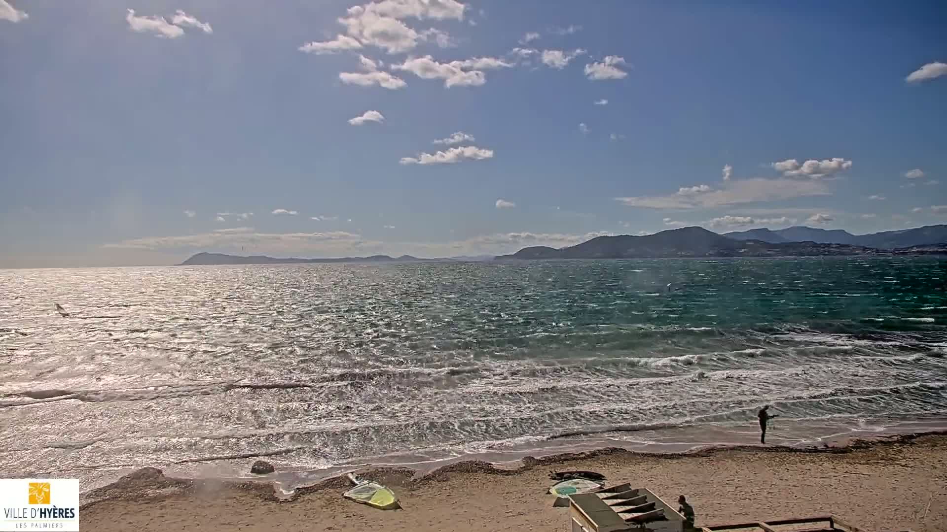 A windy, partly cloudy day at a beach with choppy water, distant mountains, and a few people on the shore.